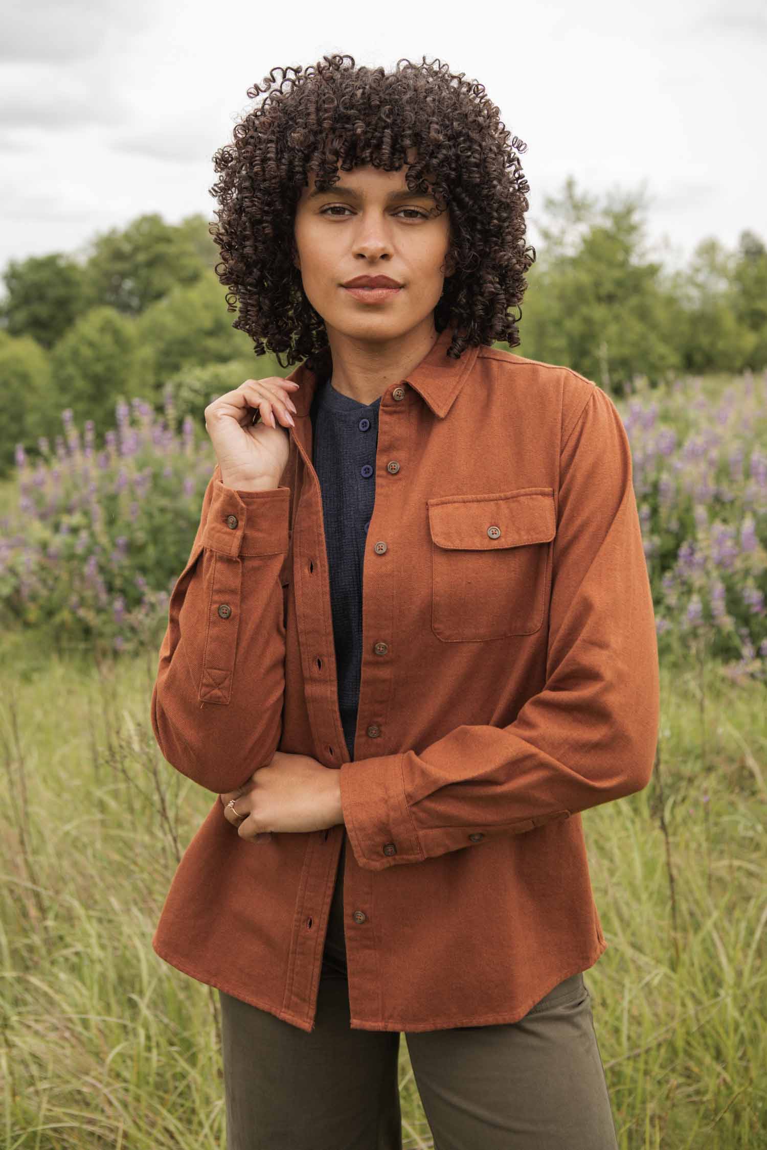 Wearing the Bridge & Burn Lenon Shirt in Terracotta Melange, a person with curly hair stands outdoors in a grassy field dotted with purple wildflowers and framed by green trees.