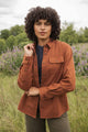 Wearing the Bridge & Burn Lenon Shirt in Terracotta Melange, a person with curly hair stands outdoors in a grassy field dotted with purple wildflowers and framed by green trees.
