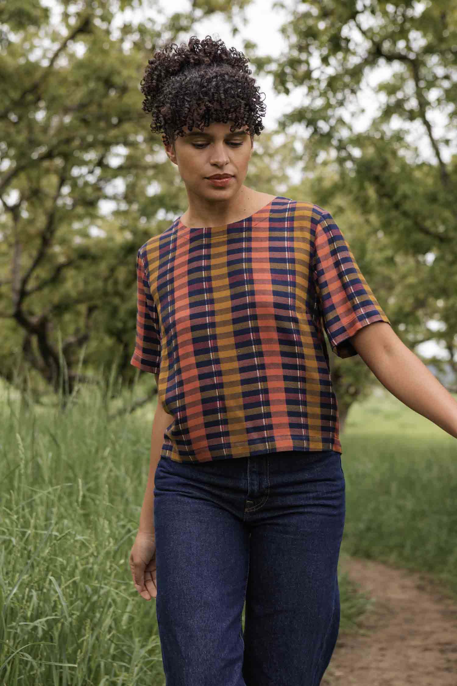 Wearing the Bridge & Burn Leona Shirt in Alpenglow Stripe, a person with curly hair and jeans walks along a grassy, tree-lined path, looking down with a calm expression.