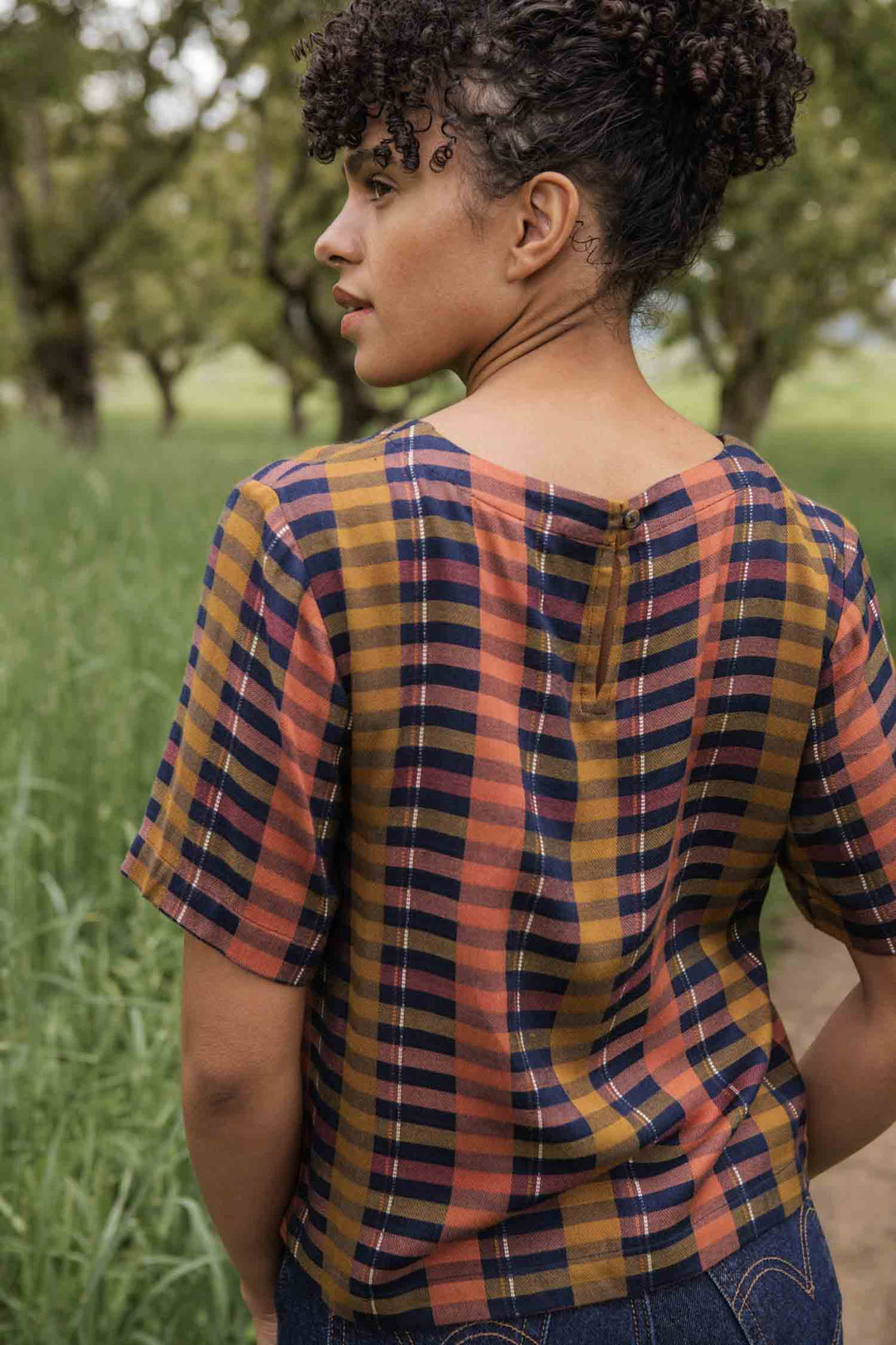 A person with curly hair stands outdoors, facing away, in the Bridge & Burn Leona Shirt / Alpenglow Stripe—a boxy fit blouse with a cropped hem—surrounded by tall grass and trees.