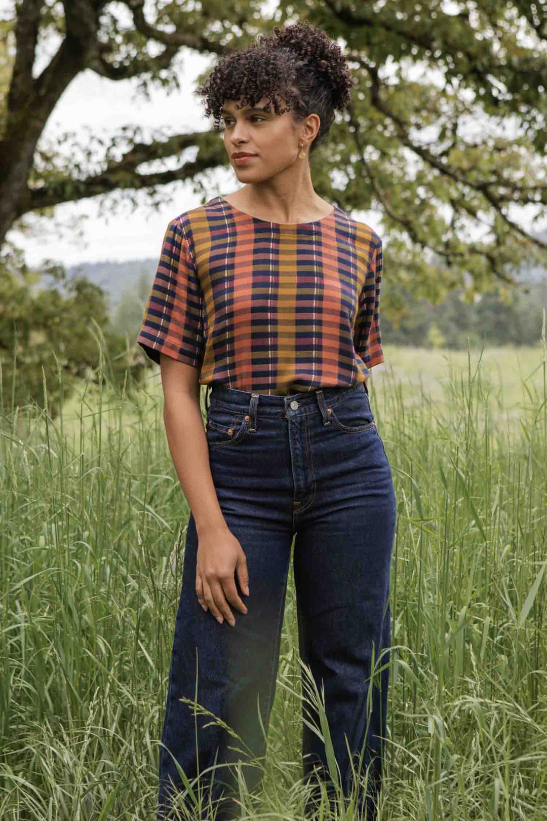 Wearing the Leona Shirt in Alpenglow Stripe by Bridge & Burn, a woman stands in tall grass and looks left. Crafted from an eco-friendly cotton alternative, her stylish, boxy-fit blouse is set against a scenic field and trees.