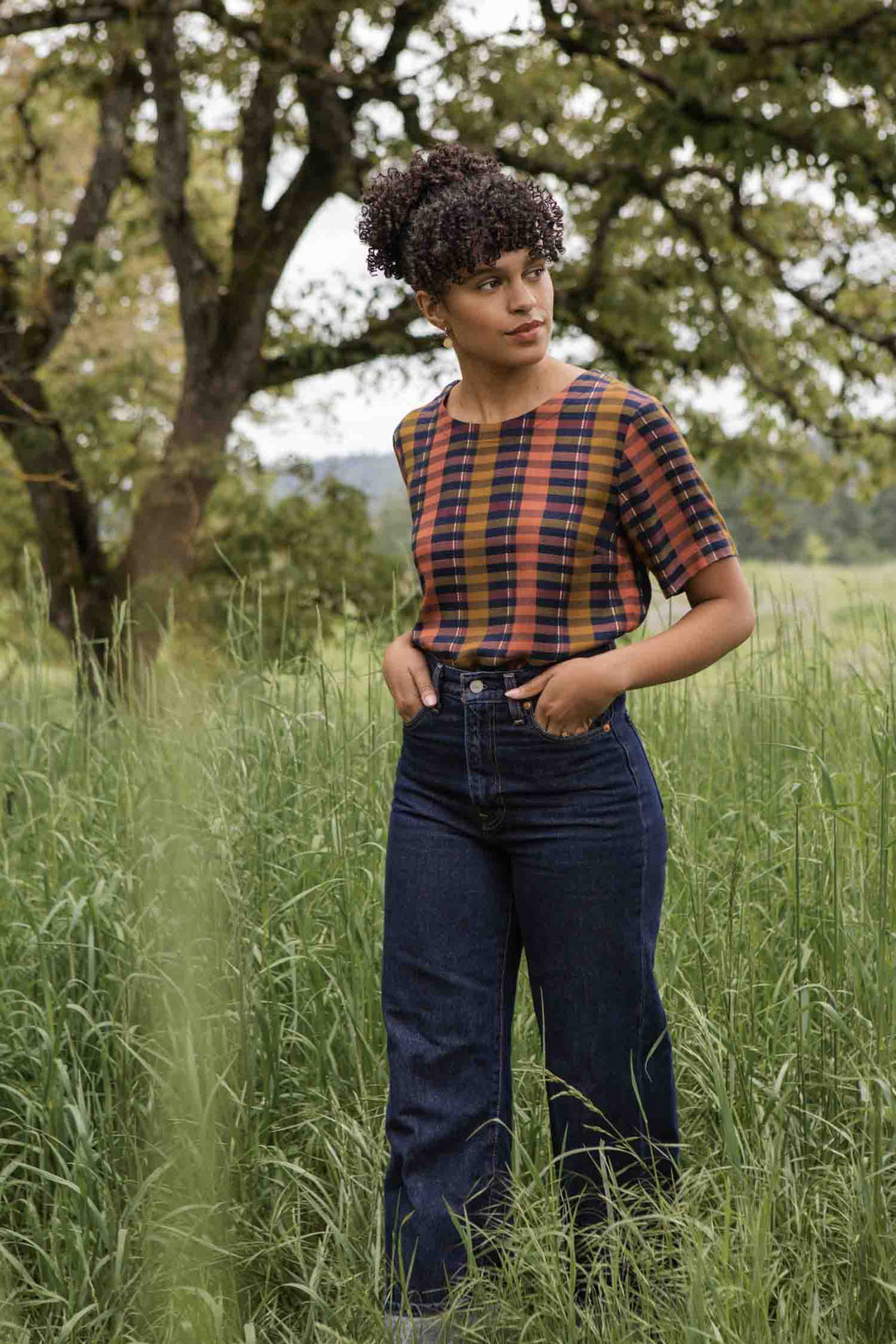 A woman with curly hair stands in tall grass, wearing the Bridge & Burn Leona Shirt in Alpenglow Stripe and high-waisted blue jeans, gazing to her left amid lush trees and greenery.