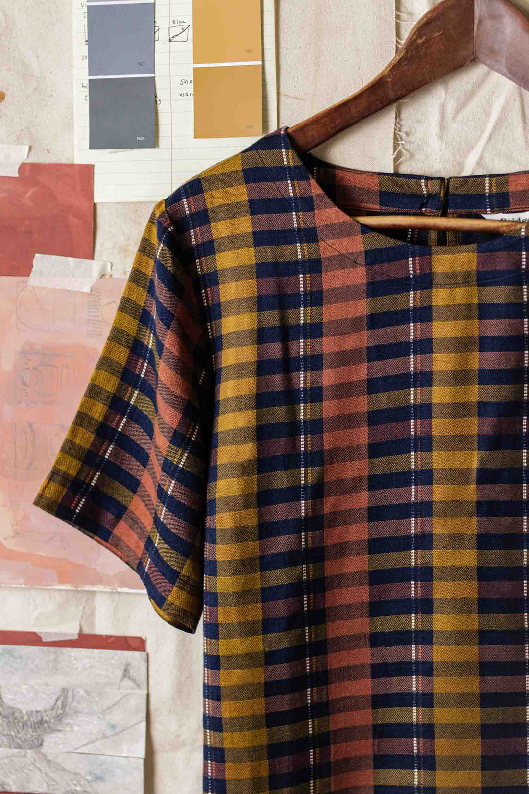 The Leona Shirt in Alpenglow Stripe by Bridge & Burn, crafted from an eco-friendly cotton alternative, features a boxy fit and multicolor plaid. It hangs on a wooden hanger against a wall with paint swatches and paper samples.