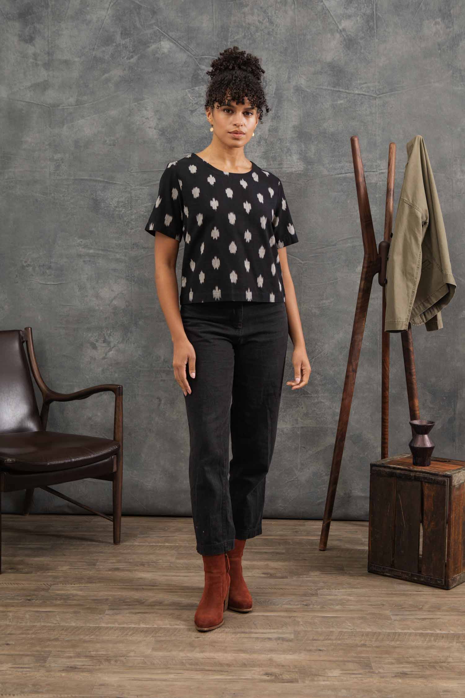 A woman stands indoors on a wooden floor, wearing the Bridge & Burn Leona Shirt in Eclipse Ikat with black pants and brown boots. Behind her are a wooden chair, coat rack with jacket, and small wooden box against a gray patterned wall.