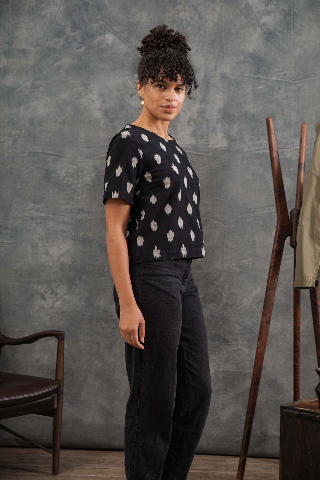 A woman with curly hair stands indoors, wearing the Bridge & Burn Leona Shirt in Eclipse Ikat and black pants. She faces slightly to the side against a gray textured background, with a chair and coat rack nearby.