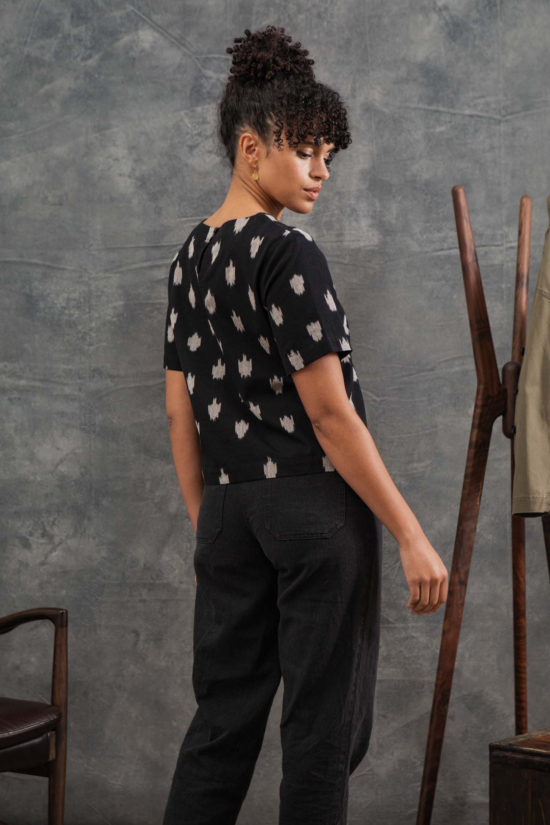 A woman with curly hair in a bun stands sideways, looking over her shoulder, wearing the Bridge & Burn Leona Shirt in Eclipse Ikat and black pants, with a wooden coat rack and a chair in the background.