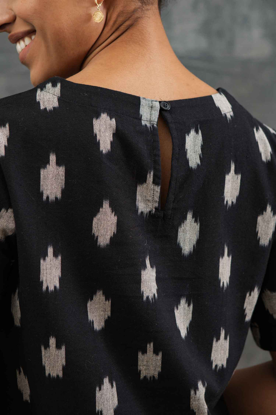Close-up of a person smiling in the Leona Shirt / Eclipse Ikat by Bridge & Burn— a black boxy-fit boatneck top featuring a white geometric pattern and single back button, complementing their short hair and small gold earring.