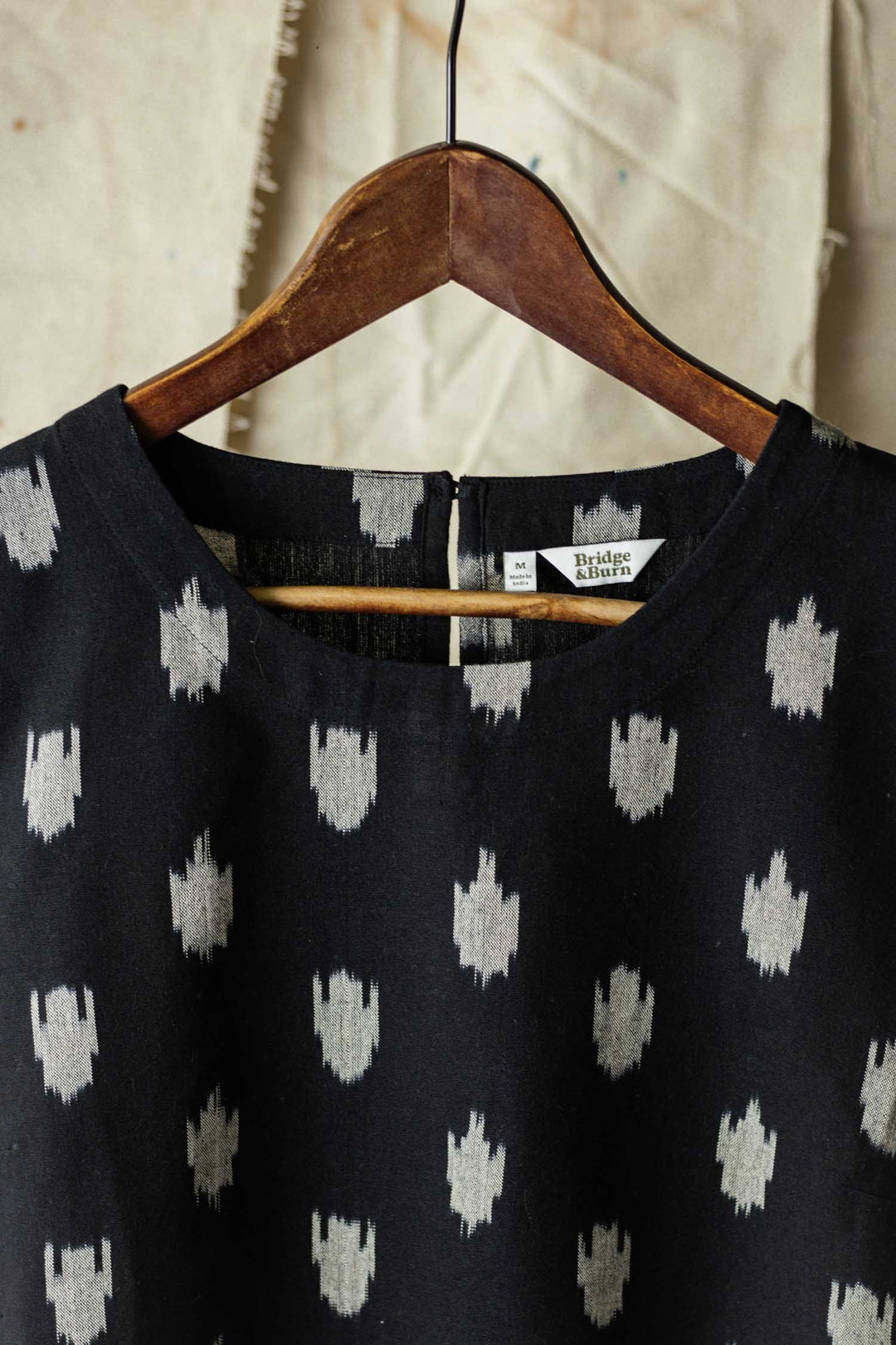 The Leona Shirt / Eclipse Ikat by Bridge & Burn hangs on a wooden hanger against a beige textured fabric background. This boxy blouse features a white abstract dot pattern, and the label reads M/L and Bridge & Burn.