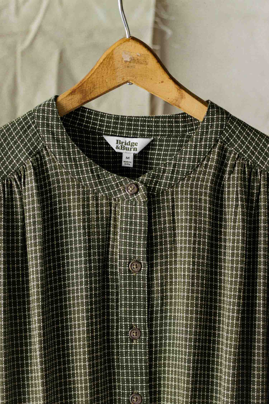 The Lorane Dress in Olive Grid Plaid by Bridge & Burn, featuring a checkered pattern and round neckline, hangs on a wooden hanger against a neutral fabric background.