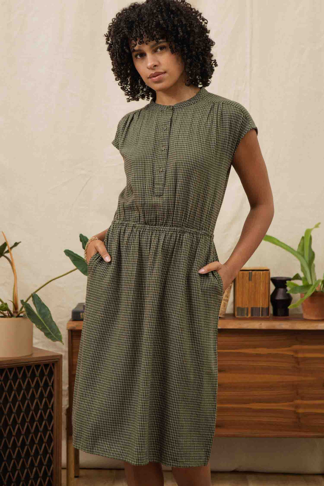 A woman with curly hair stands with her hands in the pockets of a Bridge & Burn Lorane Dress in Olive Grid Plaid, surrounded by plants and wooden furniture.