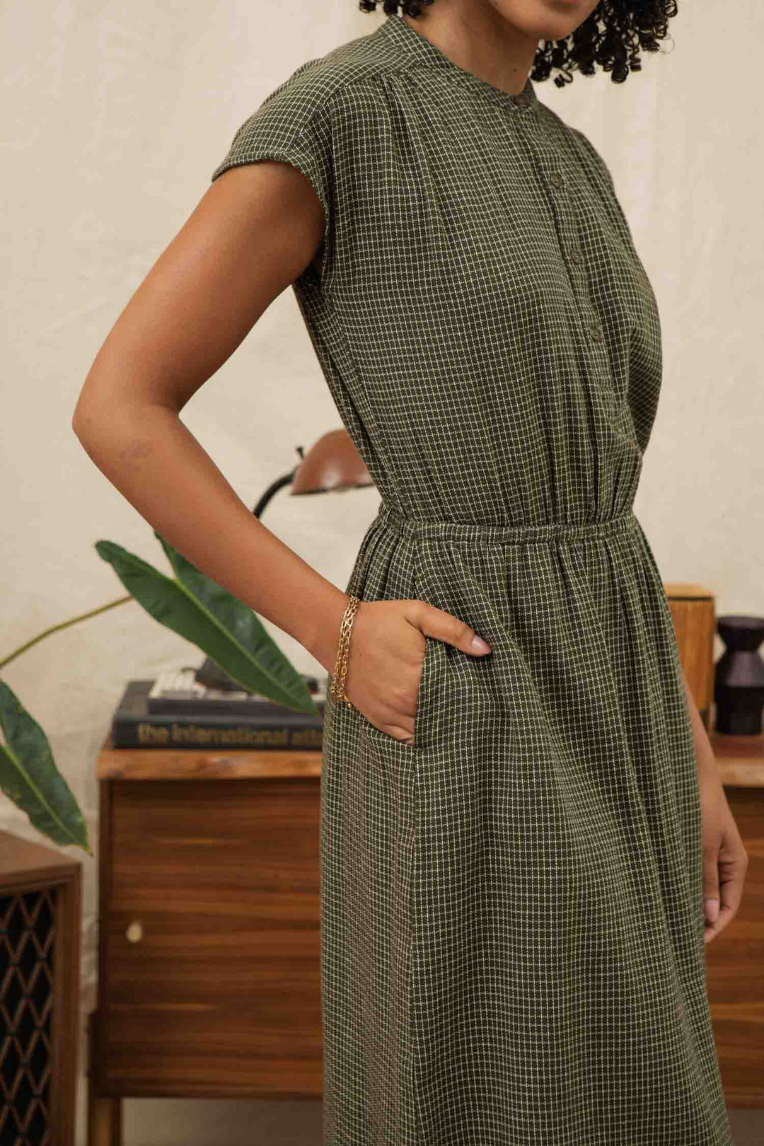 A woman with curly hair models the Bridge & Burn Lorane Dress in Olive Grid Plaid, featuring short sleeves and a cinched waist. She stands indoors with one hand in her pocket, near a wooden table holding books and a plant.