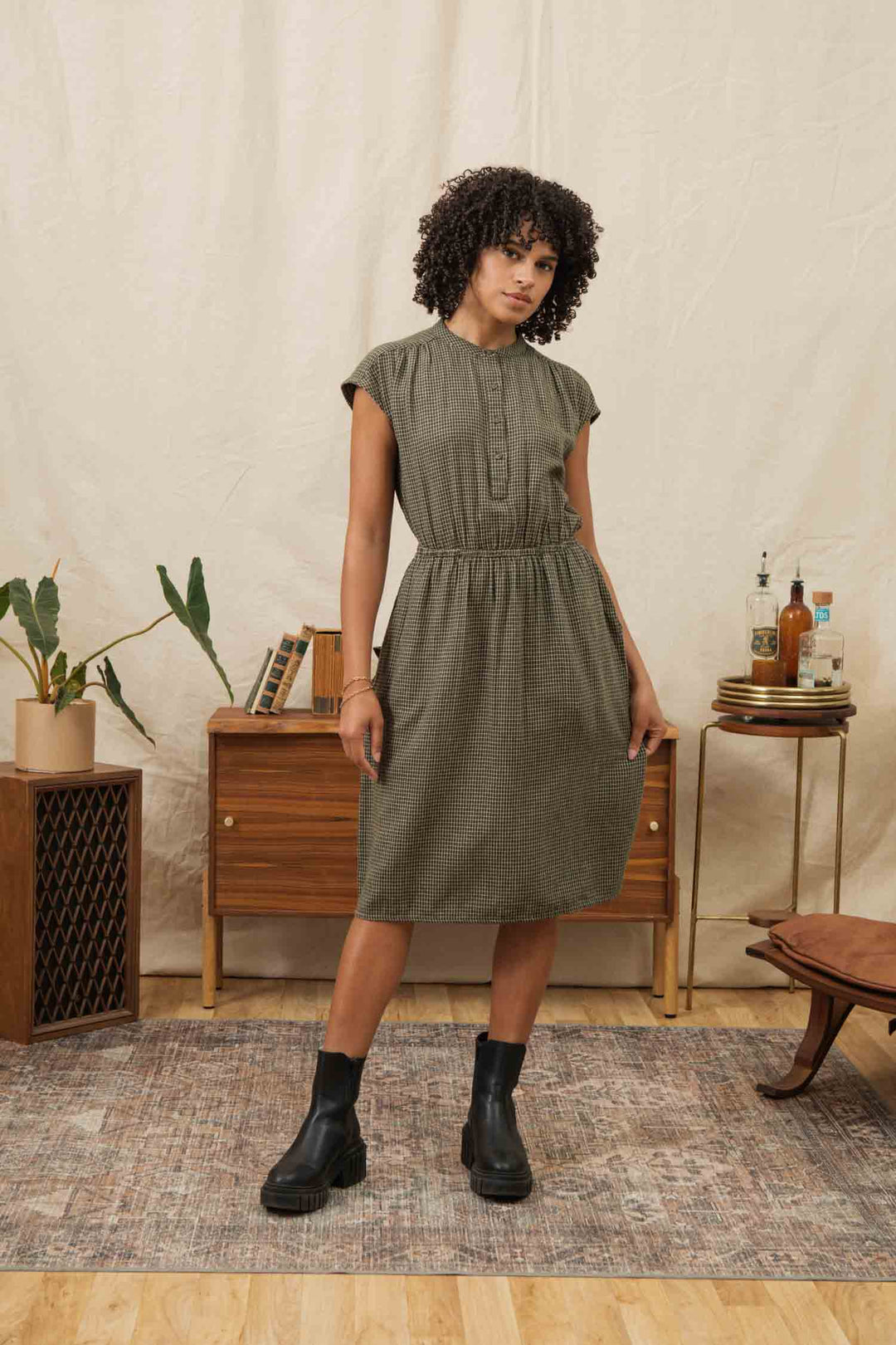 A woman with curly hair models the Bridge & Burn Lorane Dress in Olive Grid Plaid—short-sleeved, knee-length, and A-line—paired with black boots, standing indoors among wooden furniture and plants.