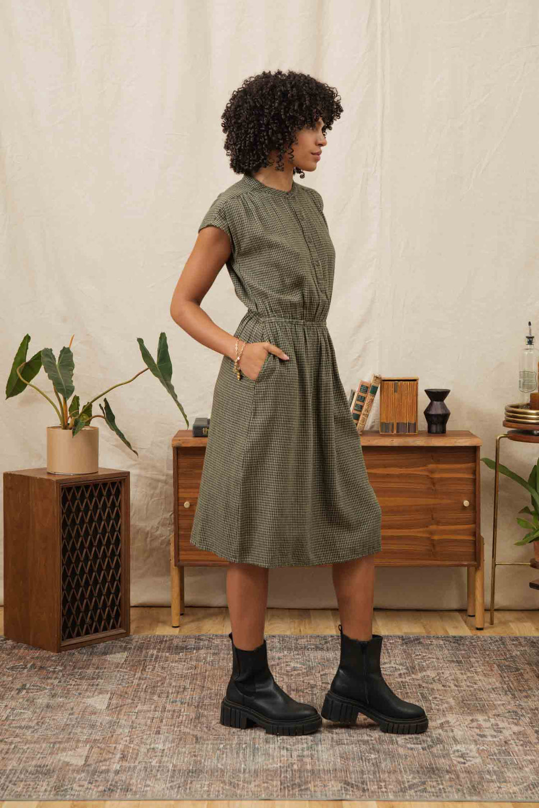 A woman with curly hair wears the Bridge & Burn Lorane Dress in Olive Grid Plaid with black boots, standing sideways with hands in pockets in a stylish room featuring plants, a wooden sideboard, and neutral decor.