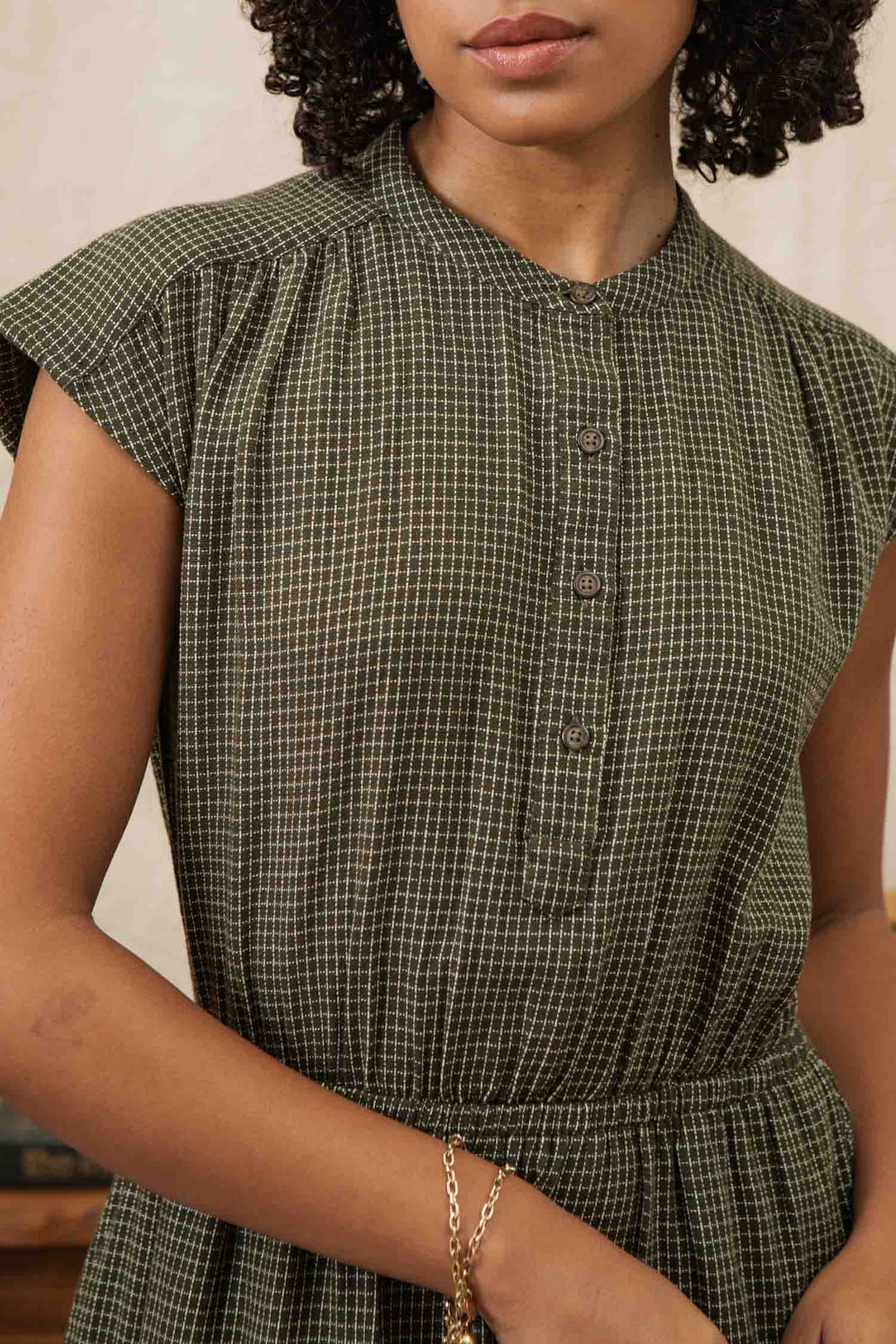 A woman wears the Bridge & Burn Lorane Dress in Olive Grid Plaid, an A-line casual dress with short sleeves, front buttons, and a gathered waist. She has curly hair, a gold bracelet, and her face is partially out of frame.