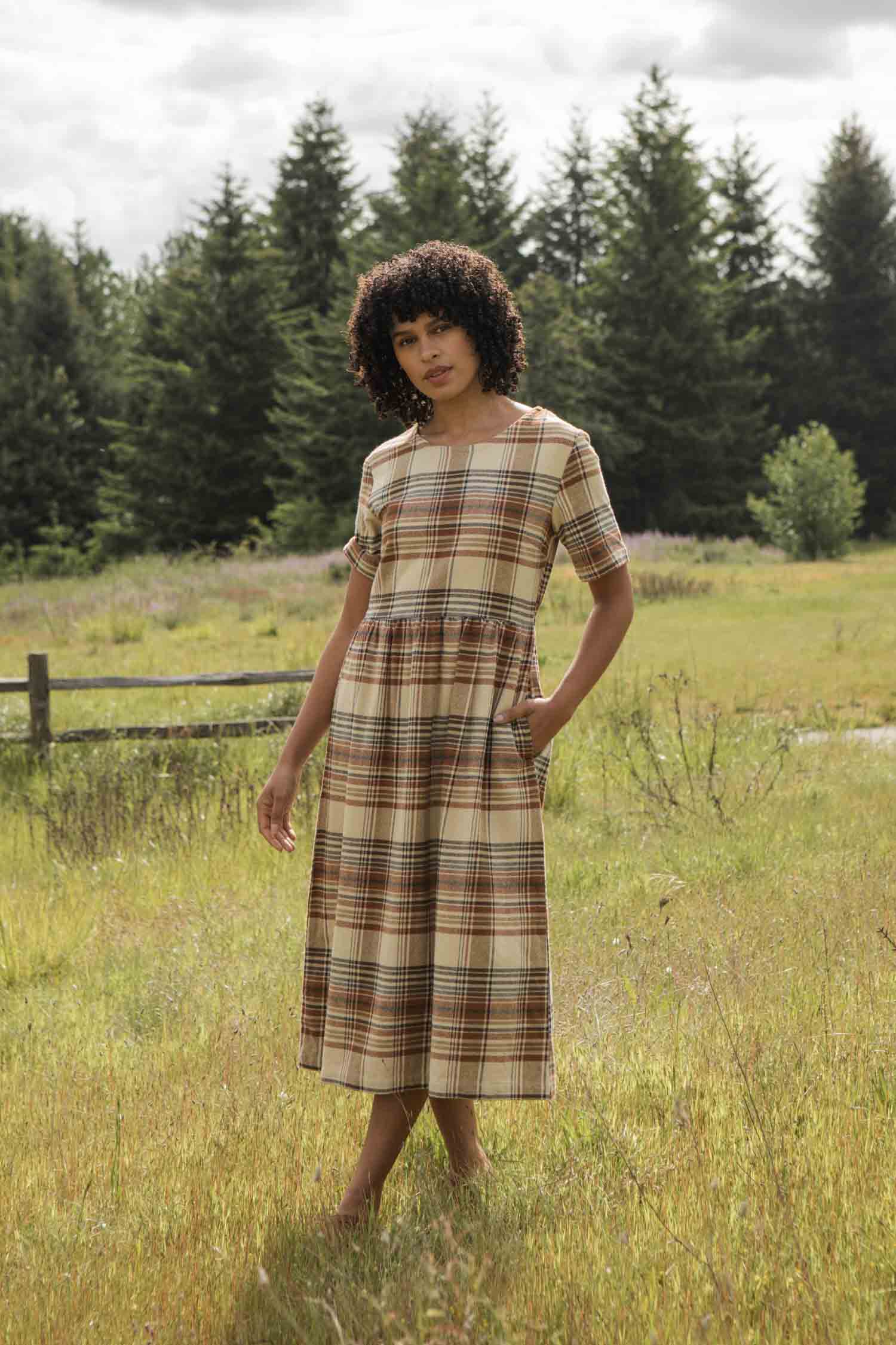 The Perry Dress / Latte Plaid by Bridge & Burn is a midi-length silhouette with short sleeves, a fitted waist, and a brown, beige, and black plaid on cream recycled cotton. A gathered skirt adds fullness for easy charm.