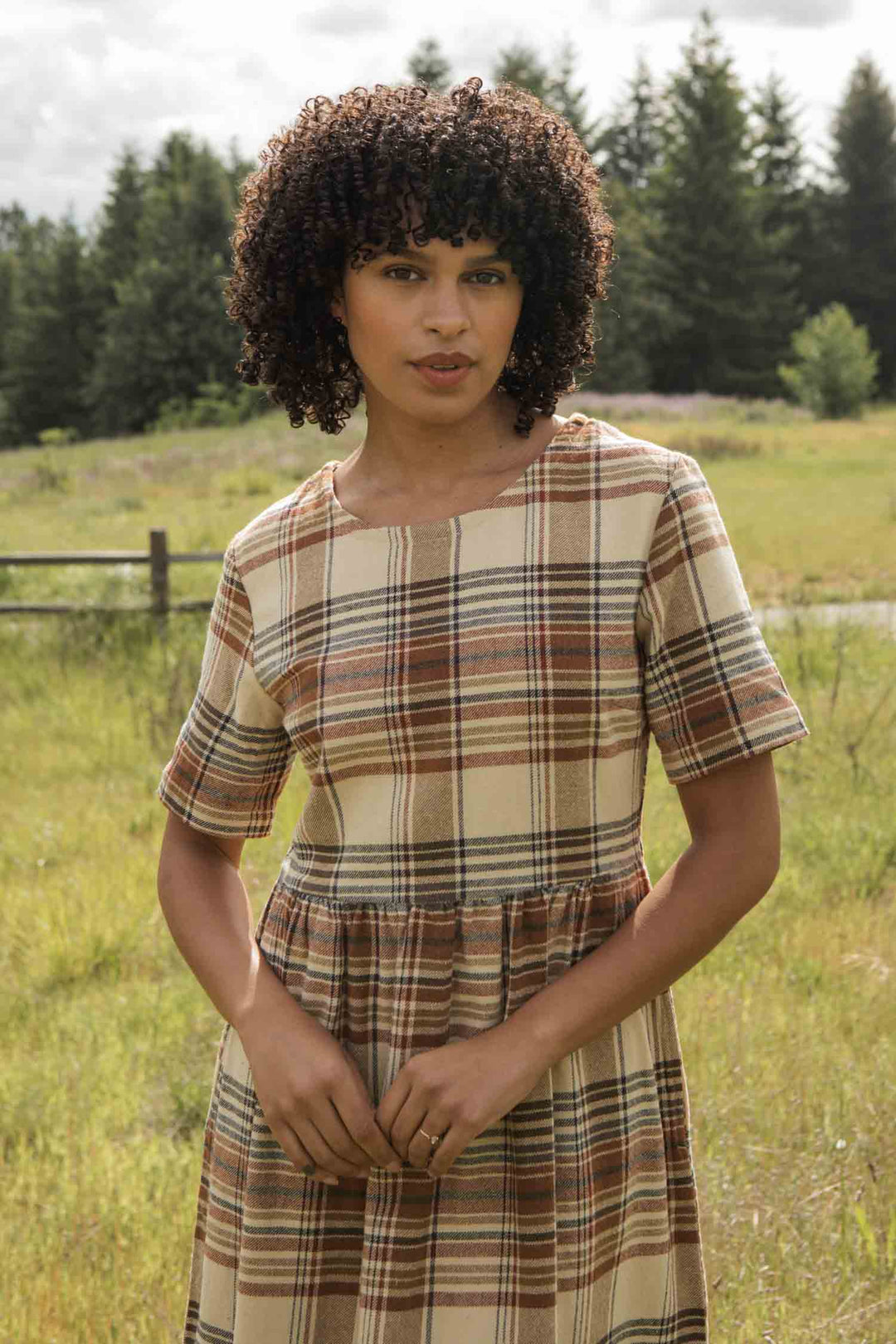 Outdoors in a grassy field, a person with curly hair wears the Bridge & Burn Perry Dress in Latte Plaid—a beige, brown, and red plaid midi dress with a gathered skirt made from recycled cotton. Trees and a wooden fence are visible behind them.