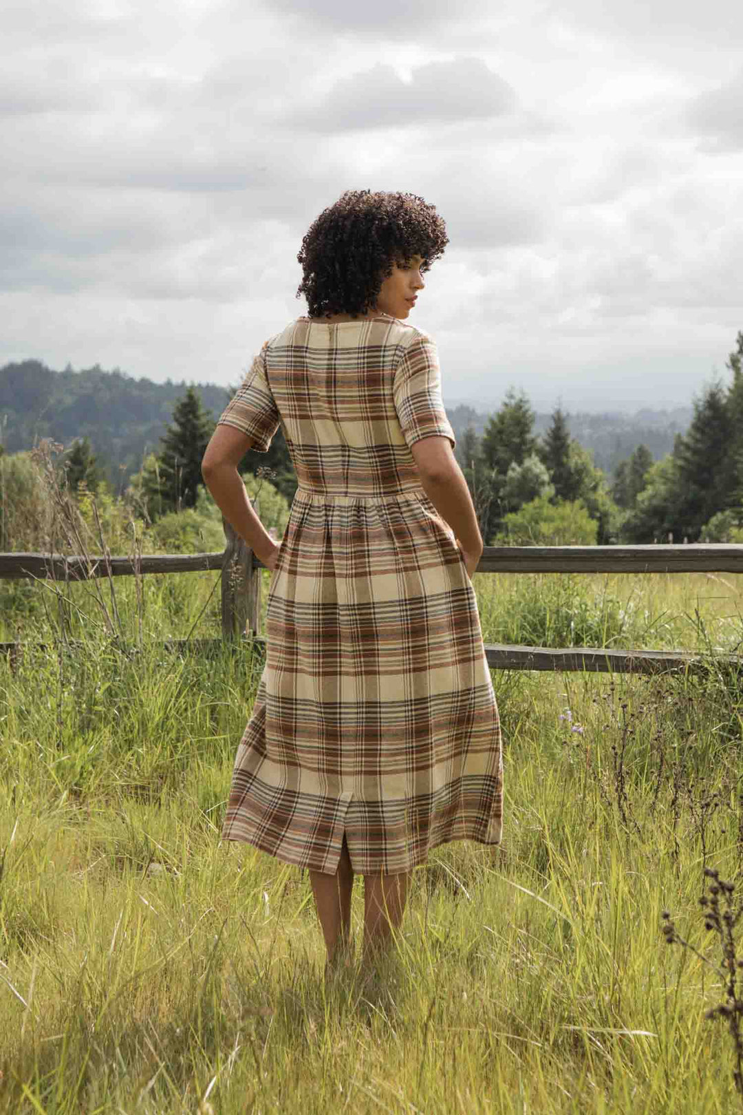 A person with curly hair stands in a grassy field, facing away from the camera, wearing the Bridge & Burn Perry Dress in Latte Plaid. Trees and rolling hills are visible under a cloudy sky.