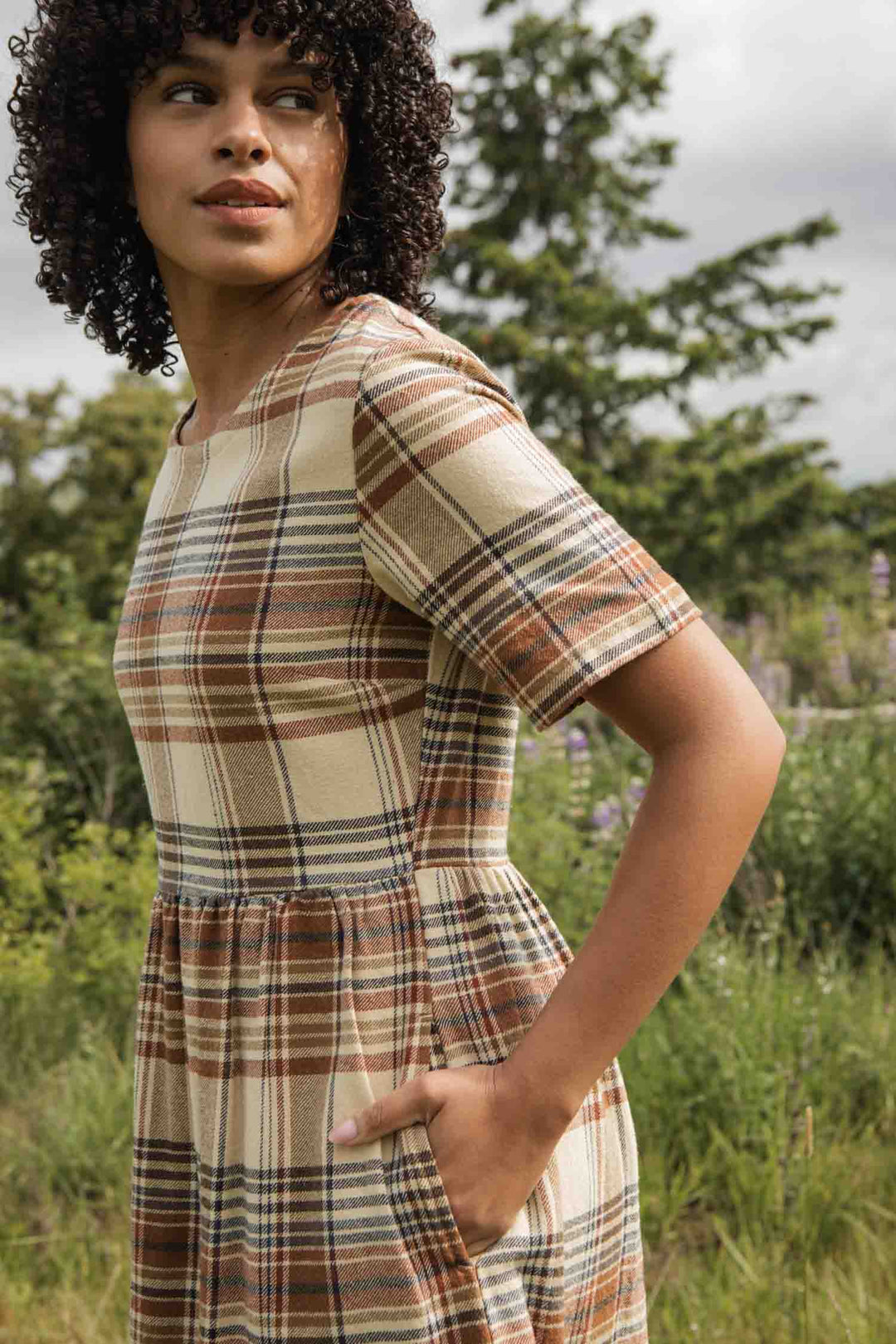 A woman with curly hair stands outdoors in greenery, wearing the Bridge & Burn Perry Dress in Latte Plaid—a beige and brown plaid midi dress with pockets and a gathered skirt. She looks to the side among trees.