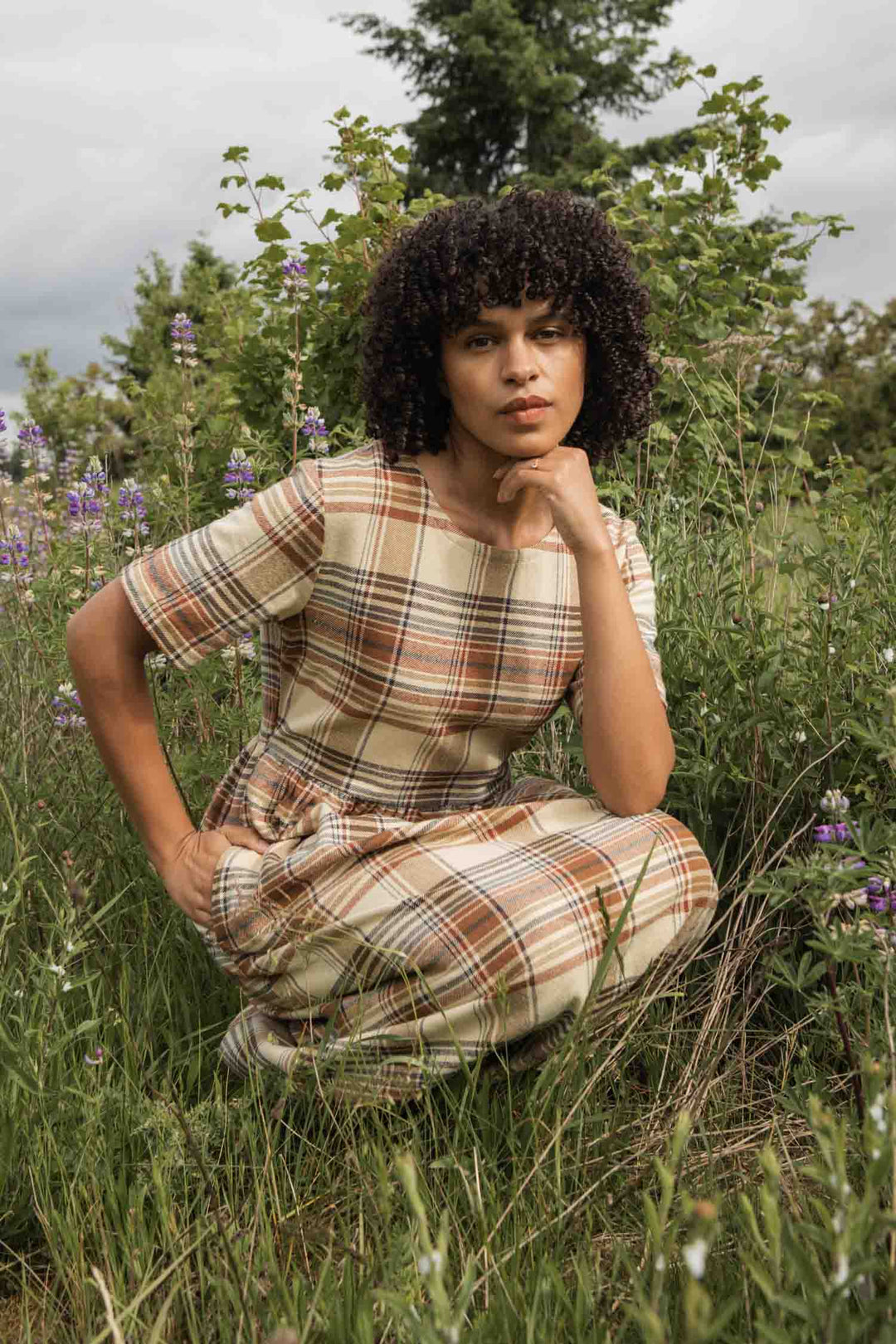 A person with curly hair wears the Bridge & Burn Perry Dress in Latte Plaid, made from recycled cotton. They squat in tall grass and wildflowers, chin resting on one hand, gazing thoughtfully at the camera with trees and a cloudy sky behind them.