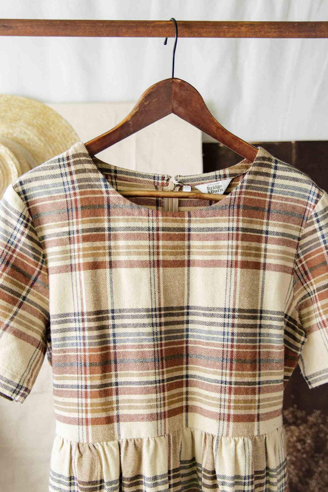 The Perry Dress / Latte Plaid by Bridge & Burn is a midi-length dress with short sleeves and a gently gathered skirt in brown, cream, and beige plaid, displayed on a wooden hanger against a soft neutral background.