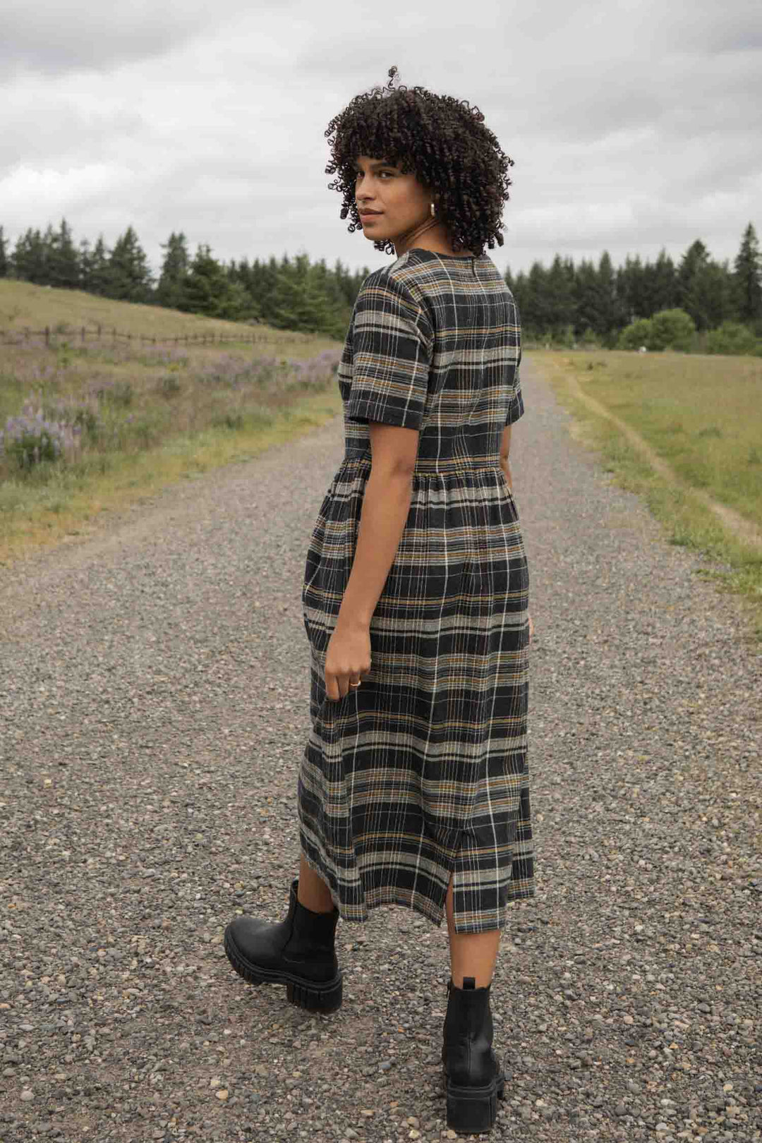 A woman with curly hair wears the Bridge & Burn Perry Dress in Volcanic Plaid with black boots, standing on a gravel path in a grassy rural area. She looks back over her shoulder under a cloudy sky, trees lining the horizon.