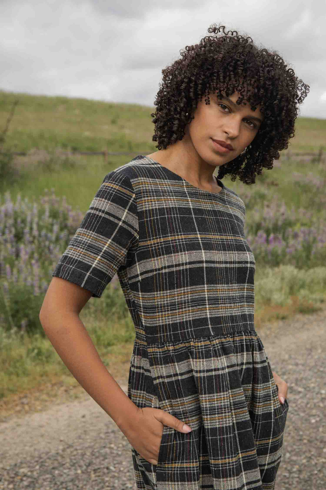 A woman with curly hair stands on a gravel path, hands in her pockets, wearing the Perry Dress in Volcanic Plaid by Bridge & Burn— a short-sleeved, midi plaid dress—against green grass and wildflowers.