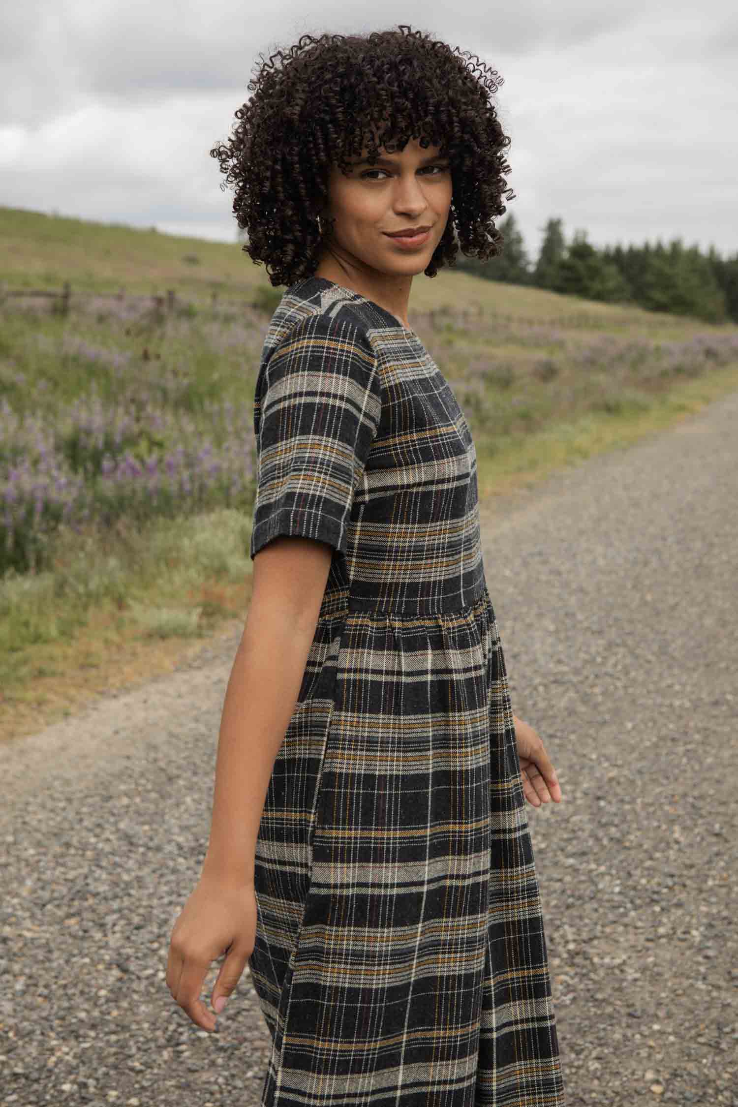 The Perry Dress in Volcanic Plaid by Bridge & Burn is a midi dress with short sleeves, made from recycled cotton. It features a dark navy, yellow, and white plaid pattern with a fitted bodice and gathered skirt.