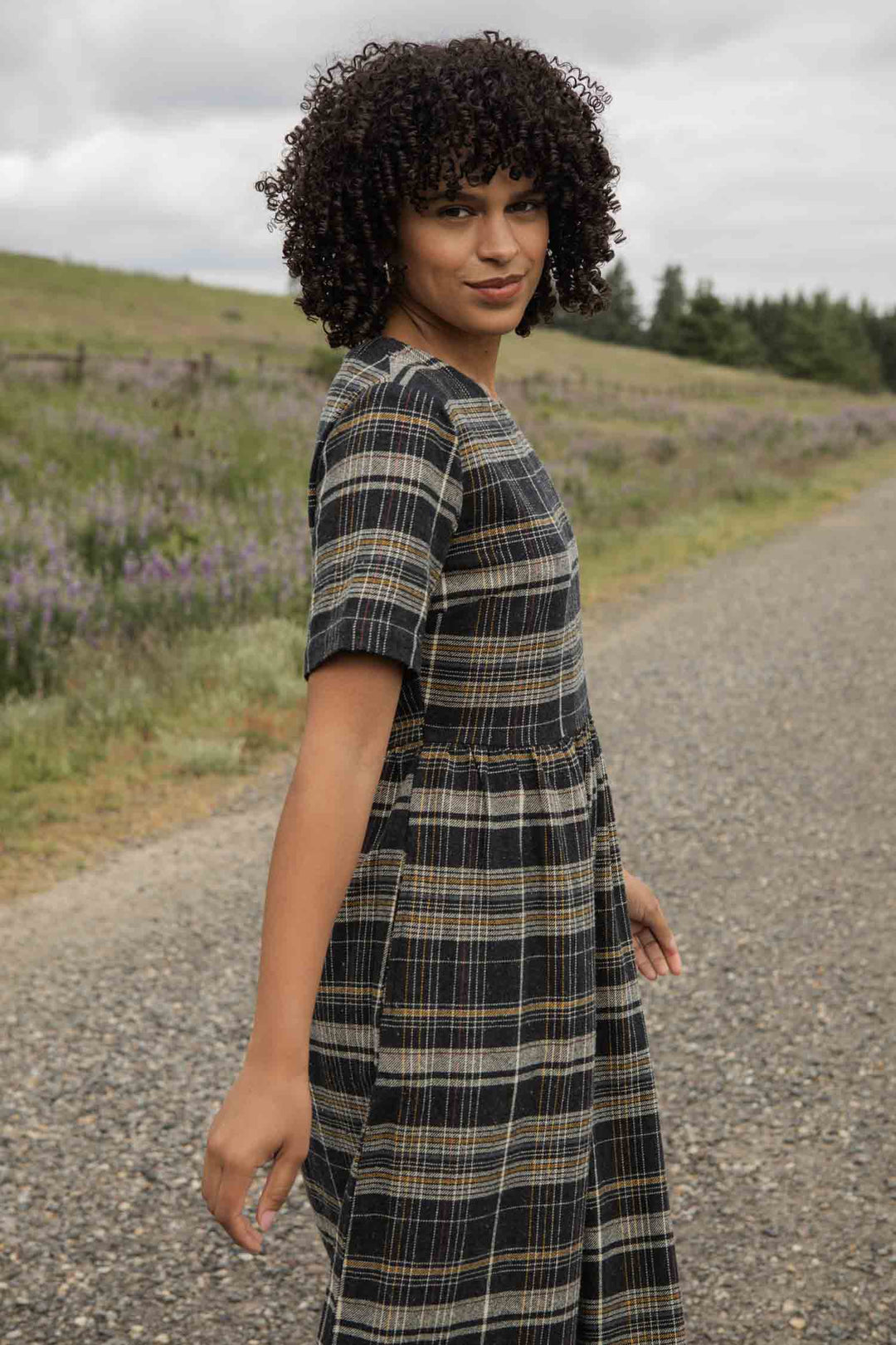 A person with curly hair stands on a gravel path in a grassy rural landscape, looking back with a slight smile while wearing the Bridge & Burn Perry Dress in Volcanic Plaid.