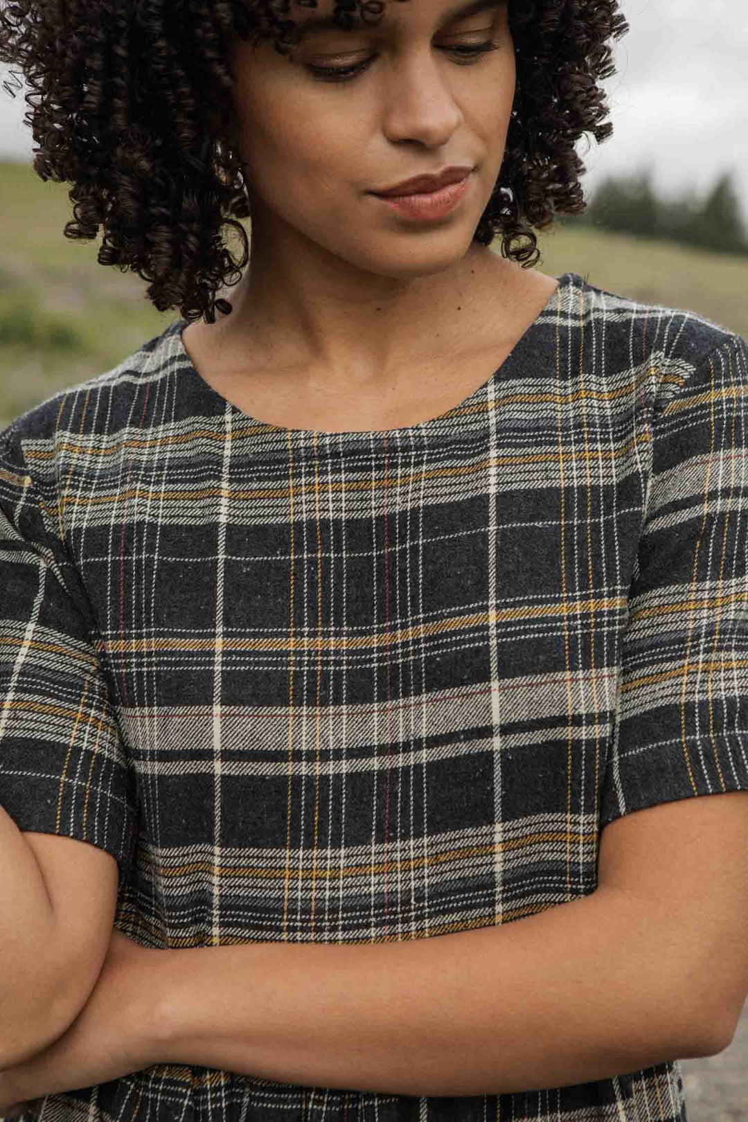 A person with curly hair stands outdoors, arms crossed and looking down, wearing Bridge & Burn’s Perry Dress in Volcanic Plaid—a short-sleeved, recycled cotton midi dress in black, gray, and yellow.