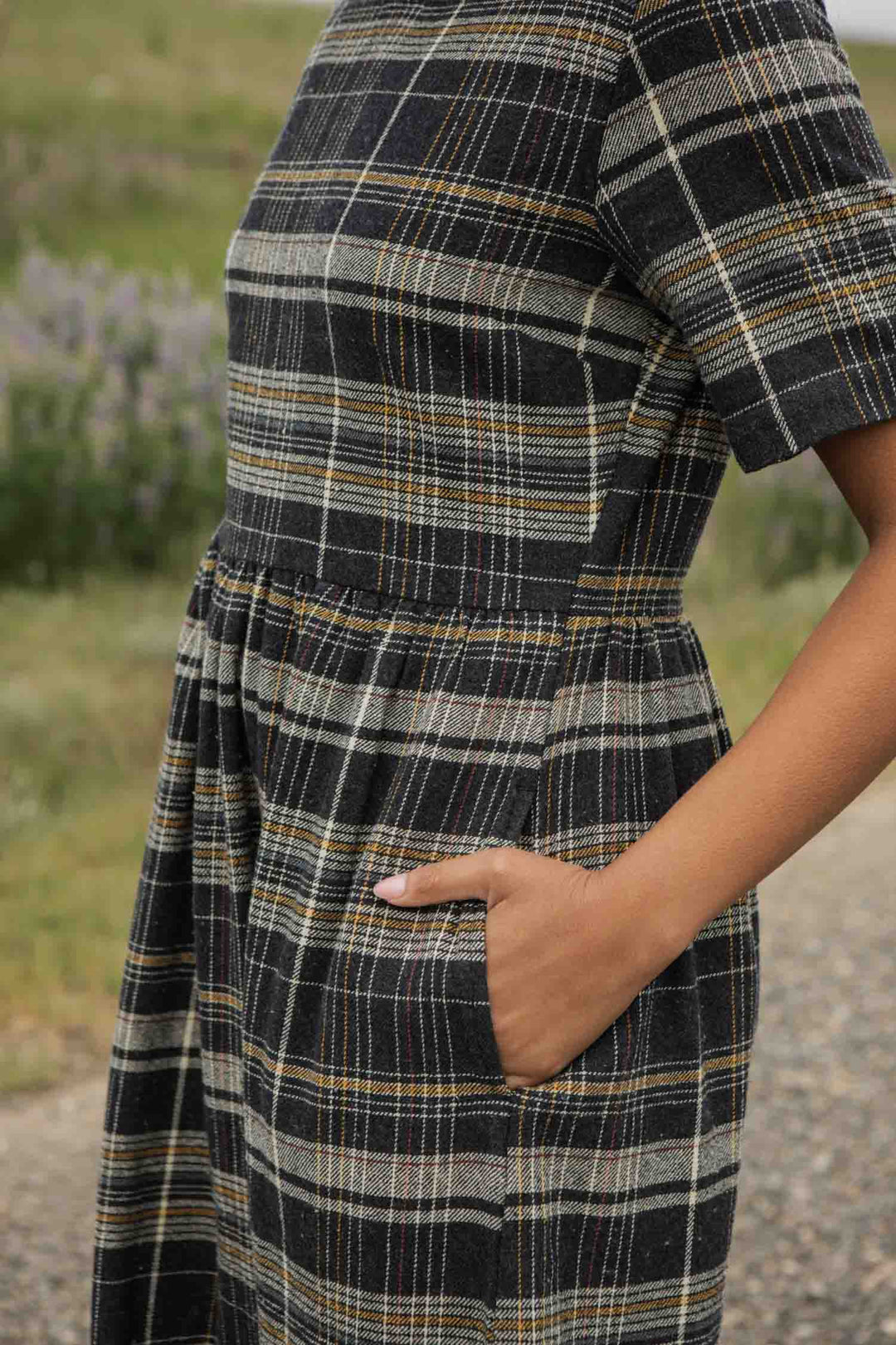 A person stands outdoors with one hand in the pocket of the Bridge & Burn Perry Dress in Volcanic Plaid—a short-sleeved, midi-length dress with a gathered skirt in gray, black, and yellow. The grassy background is slightly blurred.