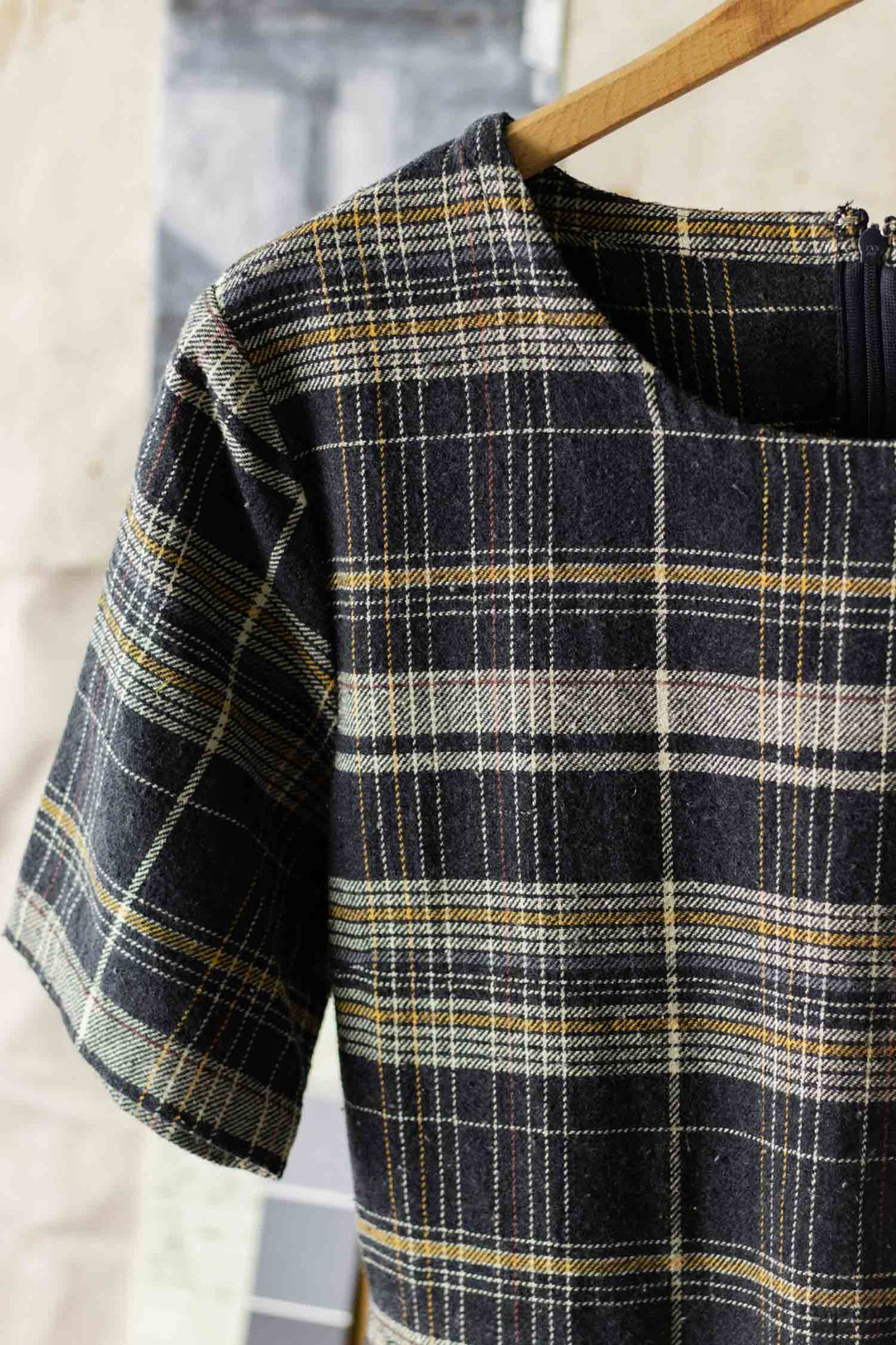 A close-up of the Bridge & Burn Perry Dress in Volcanic Plaid, a short-sleeved, midi dress with a round neckline, made from recycled cotton. The fabric features black, white, yellow, and gray intersecting lines and hangs on a wooden hanger.