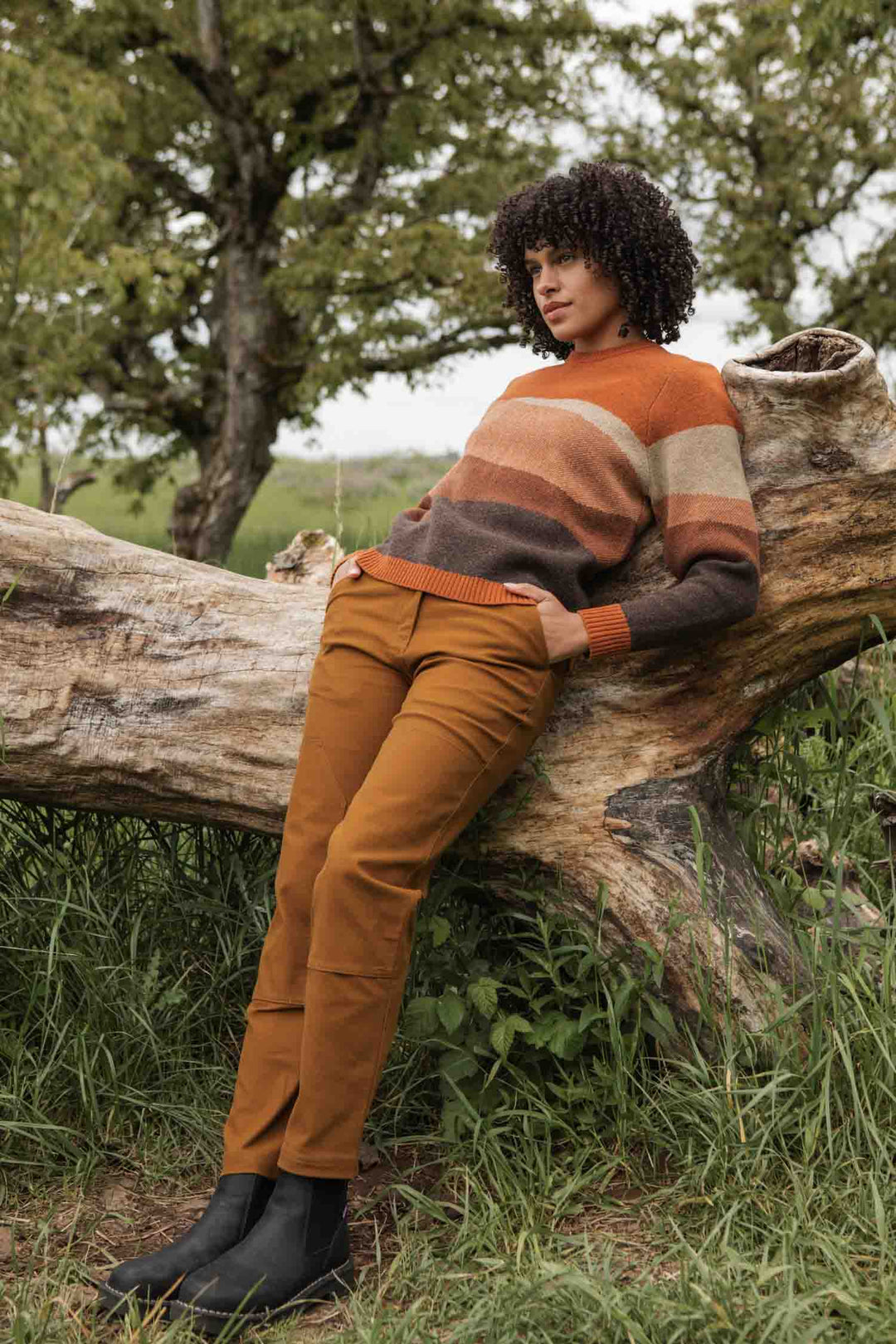 A person with curly hair leans against a fallen tree outdoors, wearing the Bridge & Burn Petra Sweater in Sandstone, rust-colored pants, and black boots, surrounded by grass and trees.