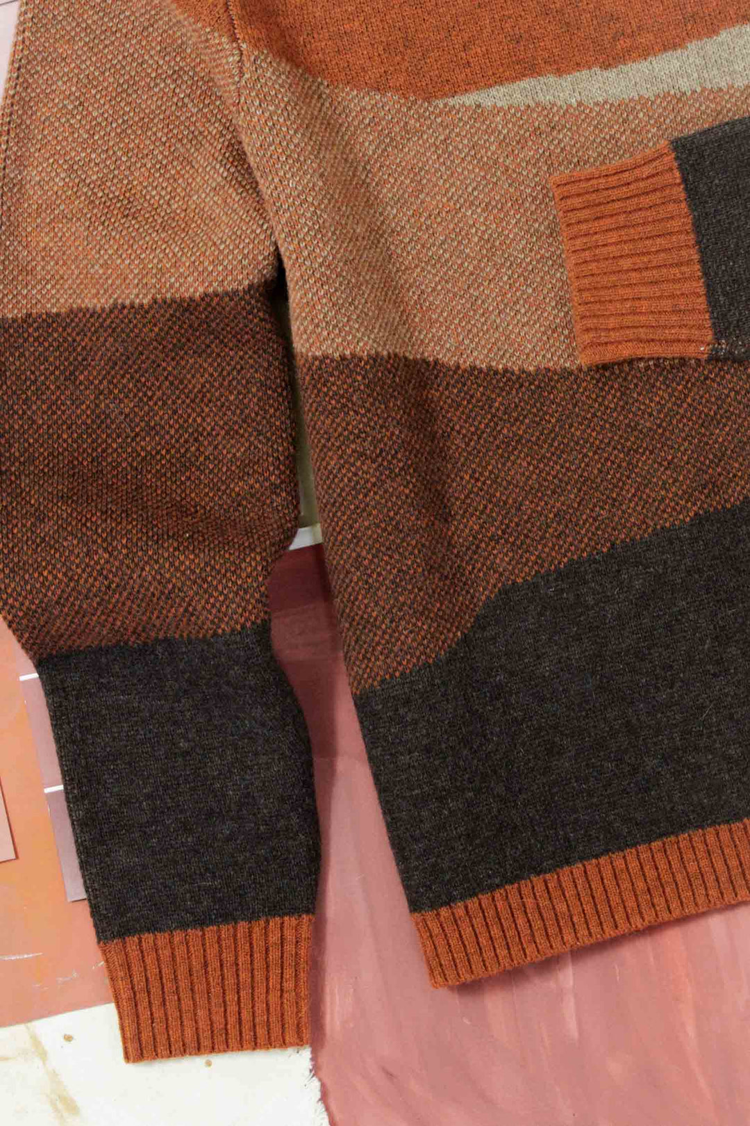A close-up of the Petra Sweater in Sandstone by Bridge & Burn, featuring wide brown, beige, and rust stripes with ribbed cuffs and hem, partially laid on a pink surface.