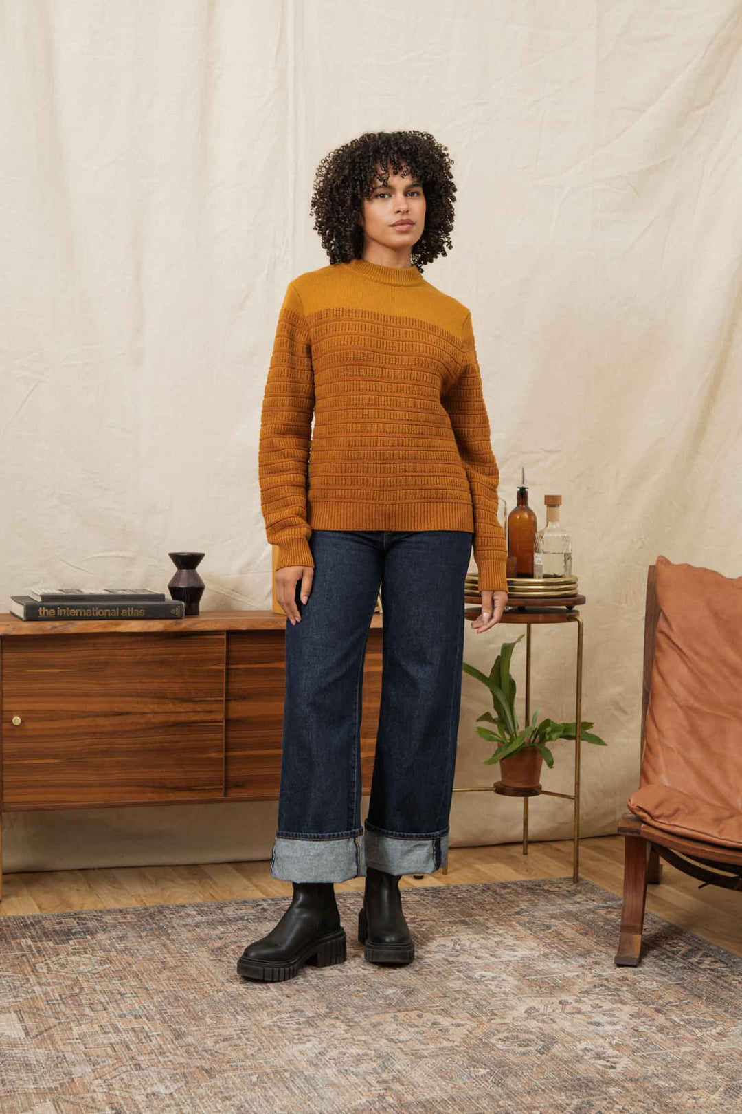 A woman with curly hair models the Bridge & Burn Sabine Sweater in Goldenrod Stripe, paired with wide cuffed jeans and black boots. She stands indoors by a wooden sideboard with books, decor, and a potted plant in a cozy, neutral-toned setting.