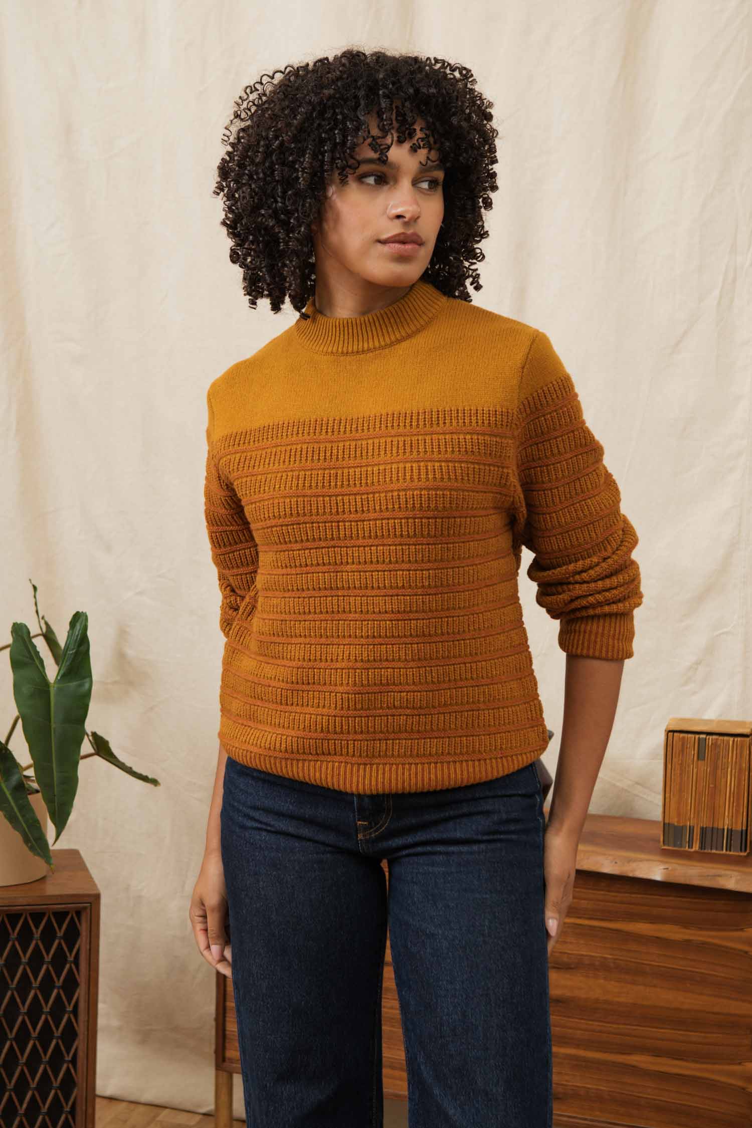 A woman with curly hair wears the Bridge & Burn Sabine Sweater in Goldenrod Stripe and dark jeans, standing indoors near wooden furniture and a plant against a light beige backdrop, looking slightly to the side with a neutral expression.