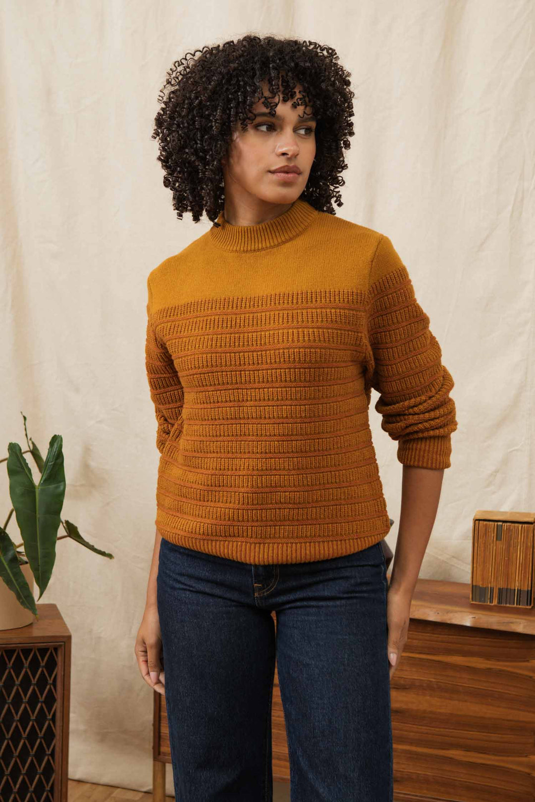 A woman with curly hair wears the Bridge & Burn Sabine Sweater in Goldenrod Stripe and dark jeans, standing indoors near wooden furniture and a plant against a light beige backdrop, looking slightly to the side with a neutral expression.