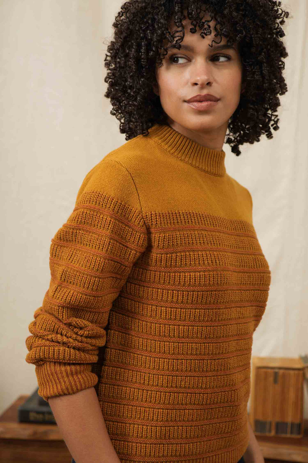A person with curly hair wears the Bridge & Burn Sabine Sweater in Goldenrod Stripe, standing indoors with a neutral expression against a softly lit, neutral-toned background.