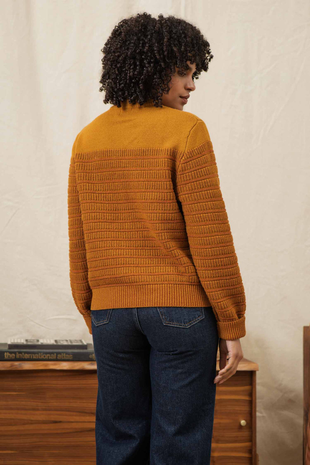 A person with curly hair, seen from behind, wears the Bridge & Burn Sabine Sweater in Goldenrod Stripe and dark jeans in a room with a wooden cabinet and books.