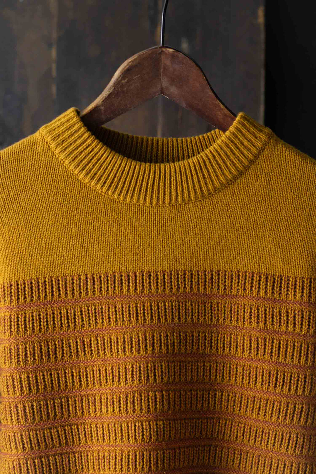 The Bridge & Burn Sabine Sweater in Goldenrod Stripe features a heavyweight knit, ribbed crew neckline, and textured horizontal stripes. Shown hanging on a wooden hanger against a dark background.