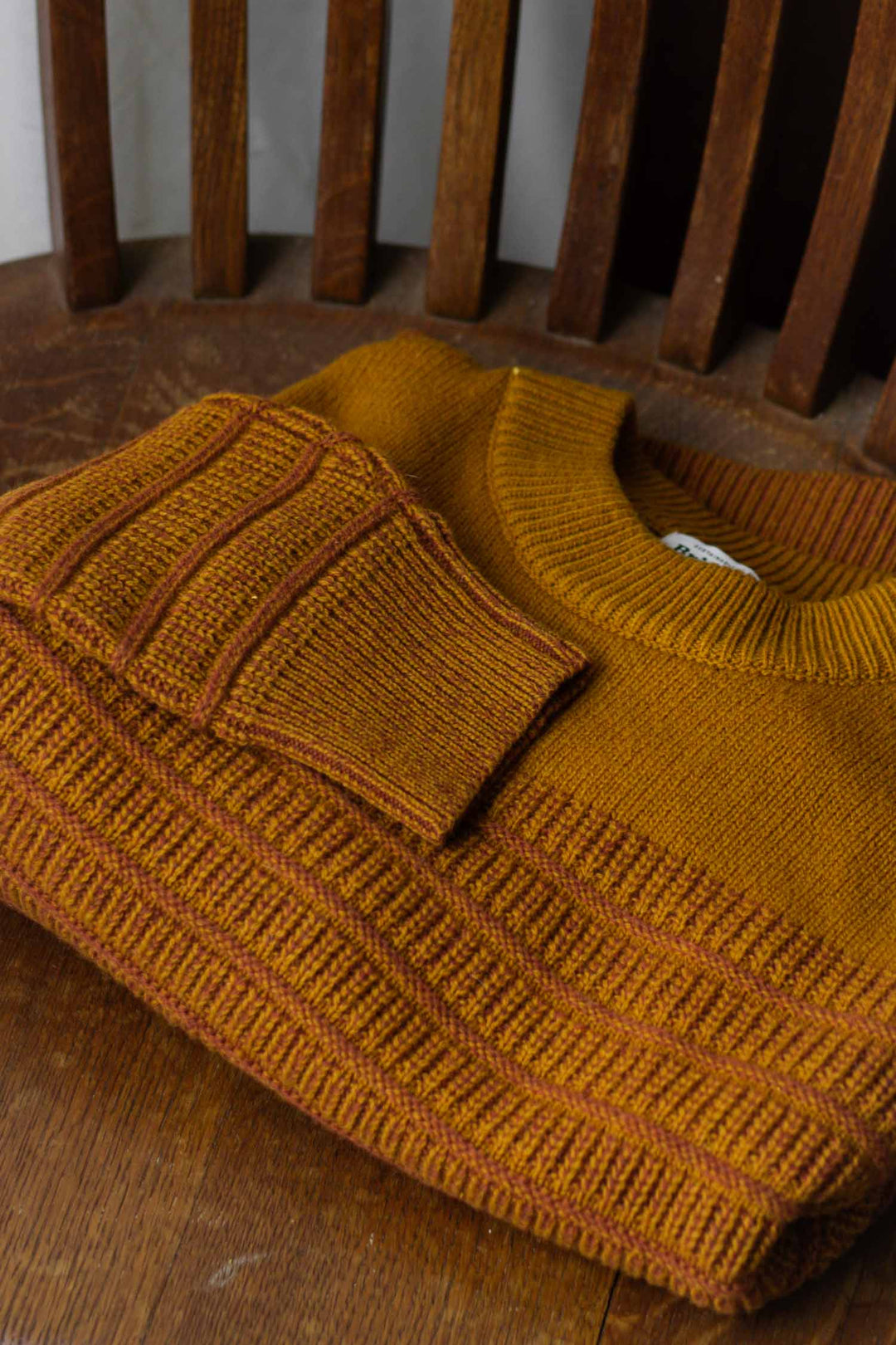 The Bridge & Burn Sabine Sweater in Goldenrod Stripe, a heavyweight knit with ribbed cuffs and hem, is folded neatly on a wooden chair with vertical slats, placed atop a wooden surface.