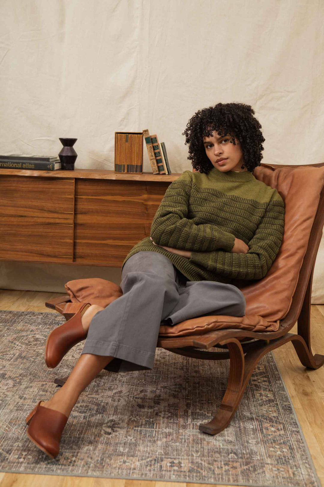 A woman with curly hair sits cross-legged on a brown chair, wearing the Bridge & Burn Sabine Sweater in olive and gray pants. She looks at the camera with a neutral expression; a wooden sideboard and books are in the background.