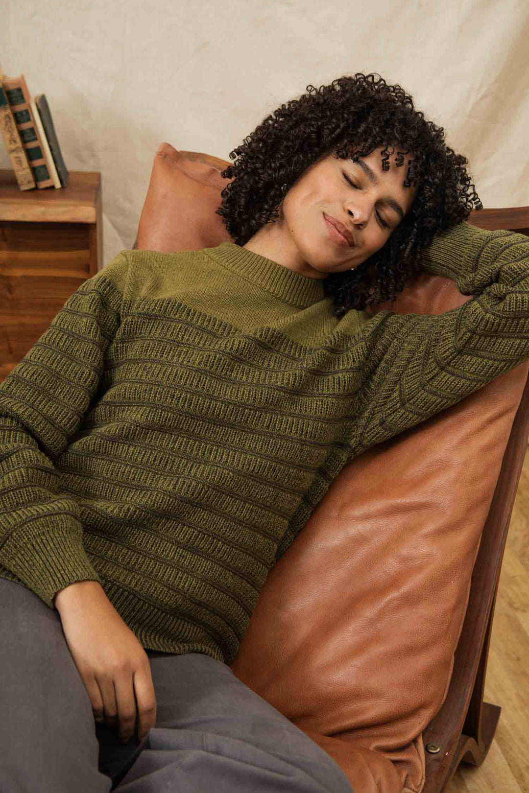 A person with curly hair reclines, eyes closed and smiling, in a brown leather chair while wearing the Bridge & Burn Sabine Sweater in Olive. A small wooden side table with books sits in the background.