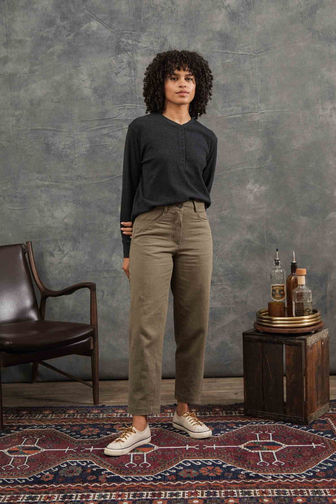 A woman with curly hair stands indoors on a patterned rug, wearing the Bridge & Burn Women's Recycled Waffle Henley in Charcoal Melange, khaki pants, and beige sneakers. She is beside a leather chair and a small table against a gray background.