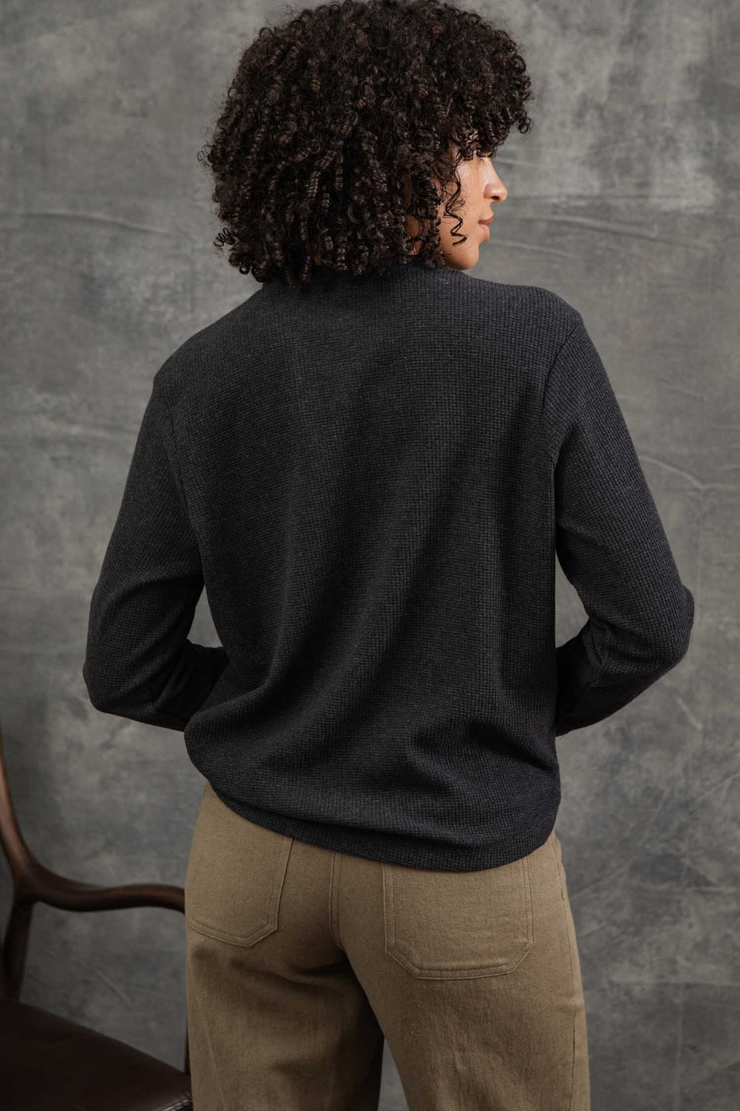 A person with curly hair models the Bridge & Burn Women's Recycled Waffle Henley in Charcoal Melange and beige recycled-fiber pants, standing with their back to the camera before a gray textured backdrop. A wooden chair appears on the left.