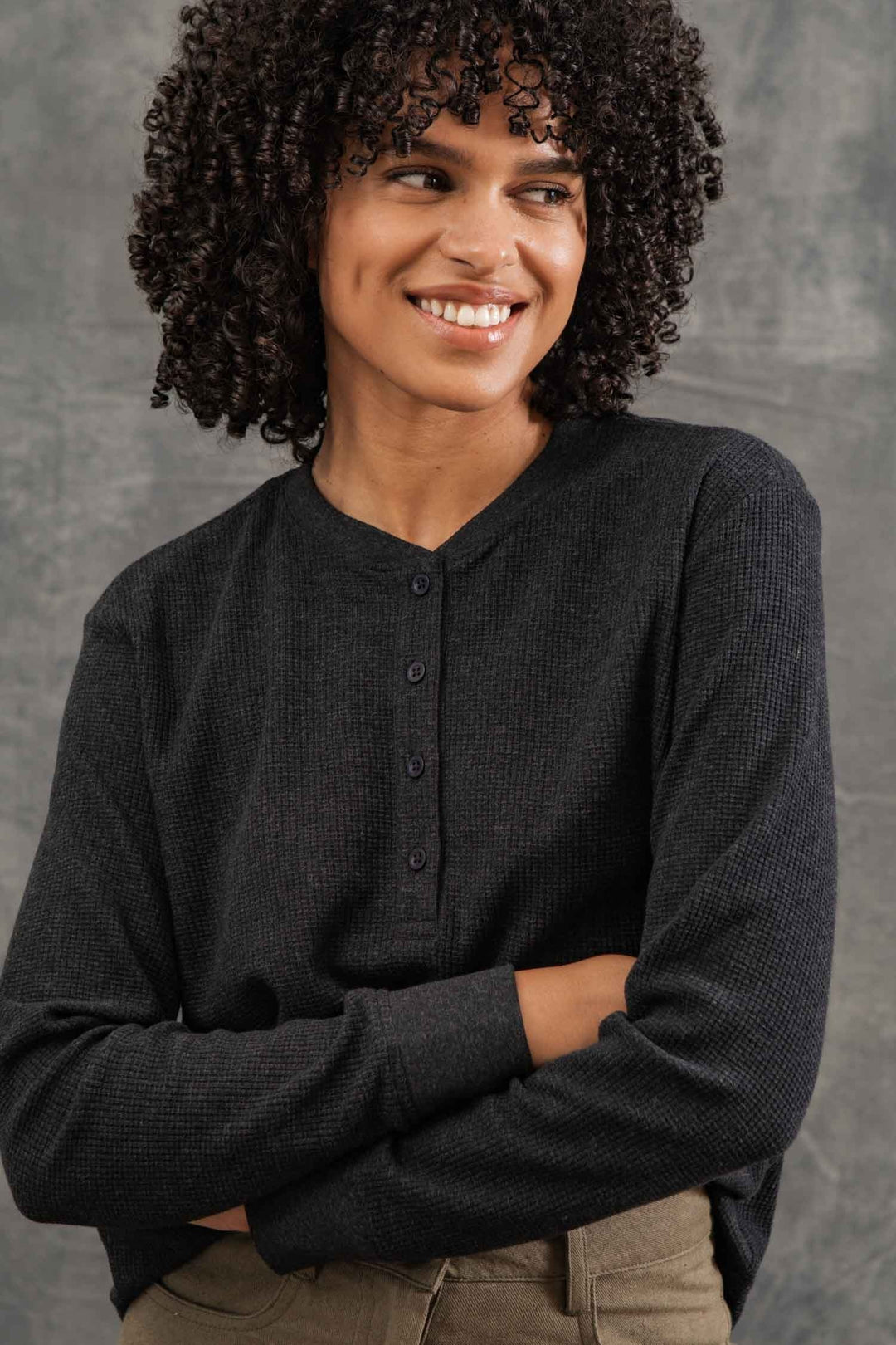 A woman with curly hair wears the Bridge & Burn Women's Recycled Waffle Henley in Charcoal Melange, standing with arms crossed and smiling while looking to the side against a gray background.