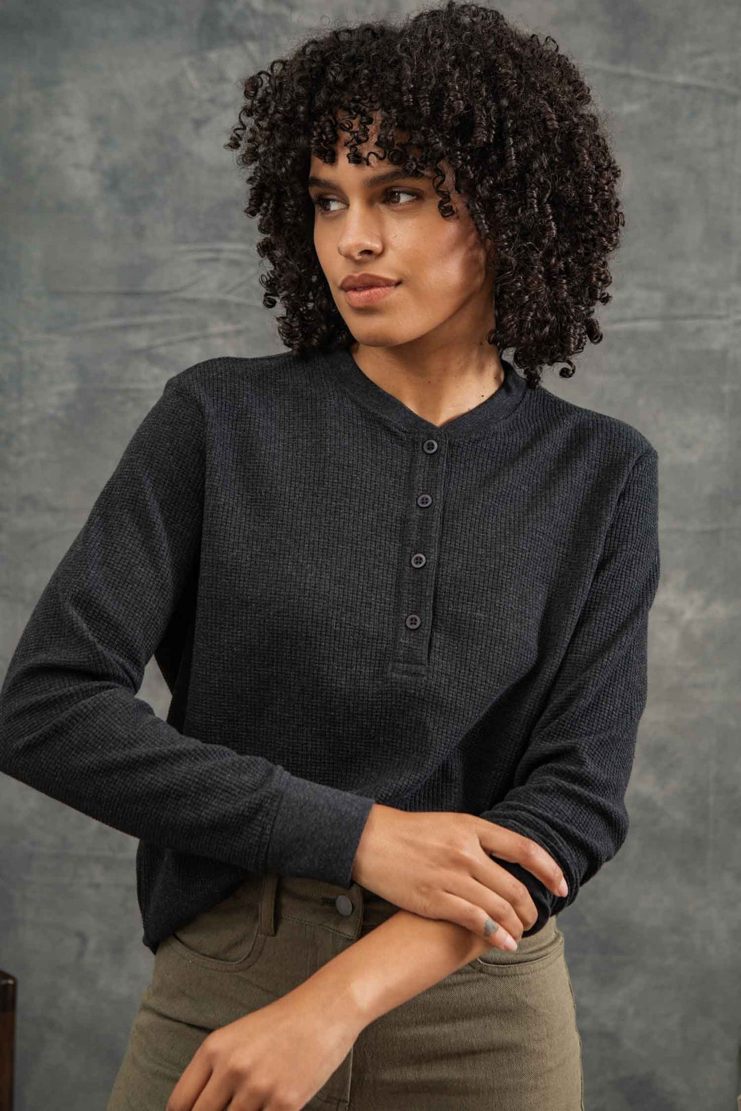 A woman with short curly hair wears the Bridge & Burn Women's Recycled Waffle Henley in Charcoal Melange and olive pants, standing before a textured gray backdrop and looking to the side with a neutral expression.