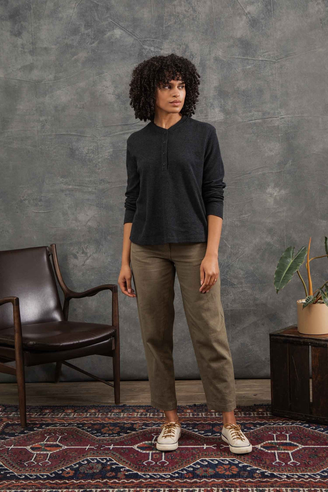 A woman with curly hair stands indoors on a patterned rug, wearing Bridge & Burn’s Women's Recycled Waffle Henley in Charcoal Melange, khaki pants, and beige sneakers. Behind her are a brown chair, wooden cabinet, and potted plant against a gray backdrop.