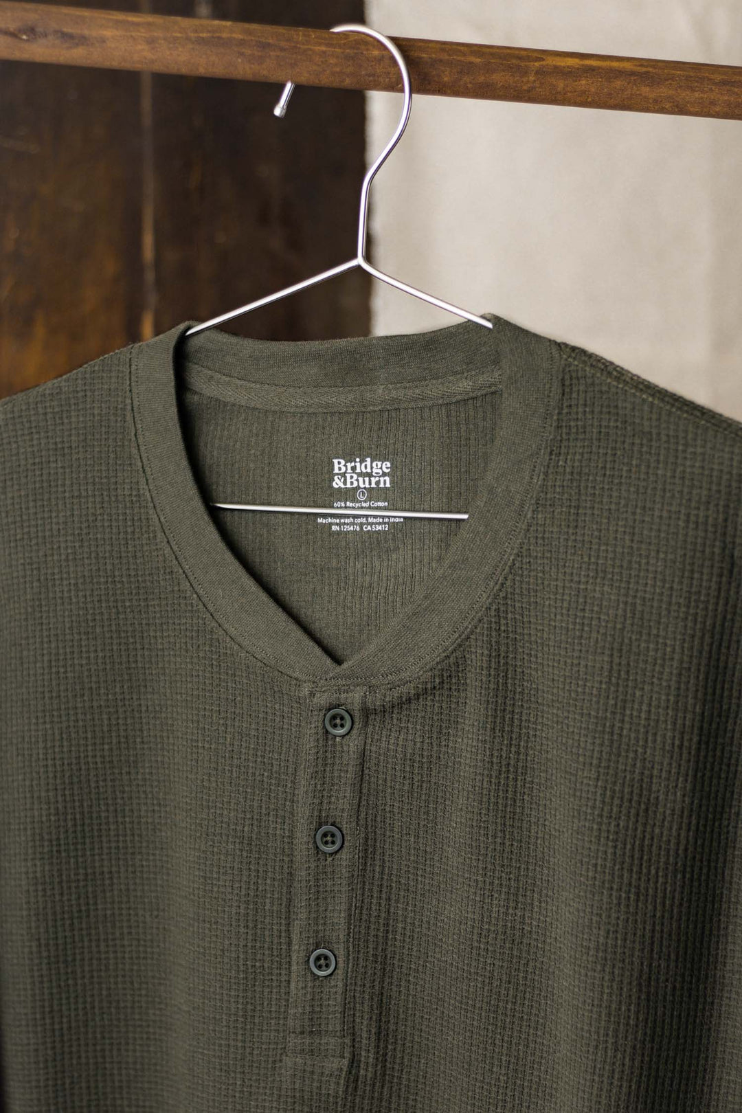 A women's forest melange, long-sleeve recycled waffle henley from Bridge & Burn with three buttons hangs on a metal hanger against a wooden background. The label displays "Bridge & Burn" in white letters.