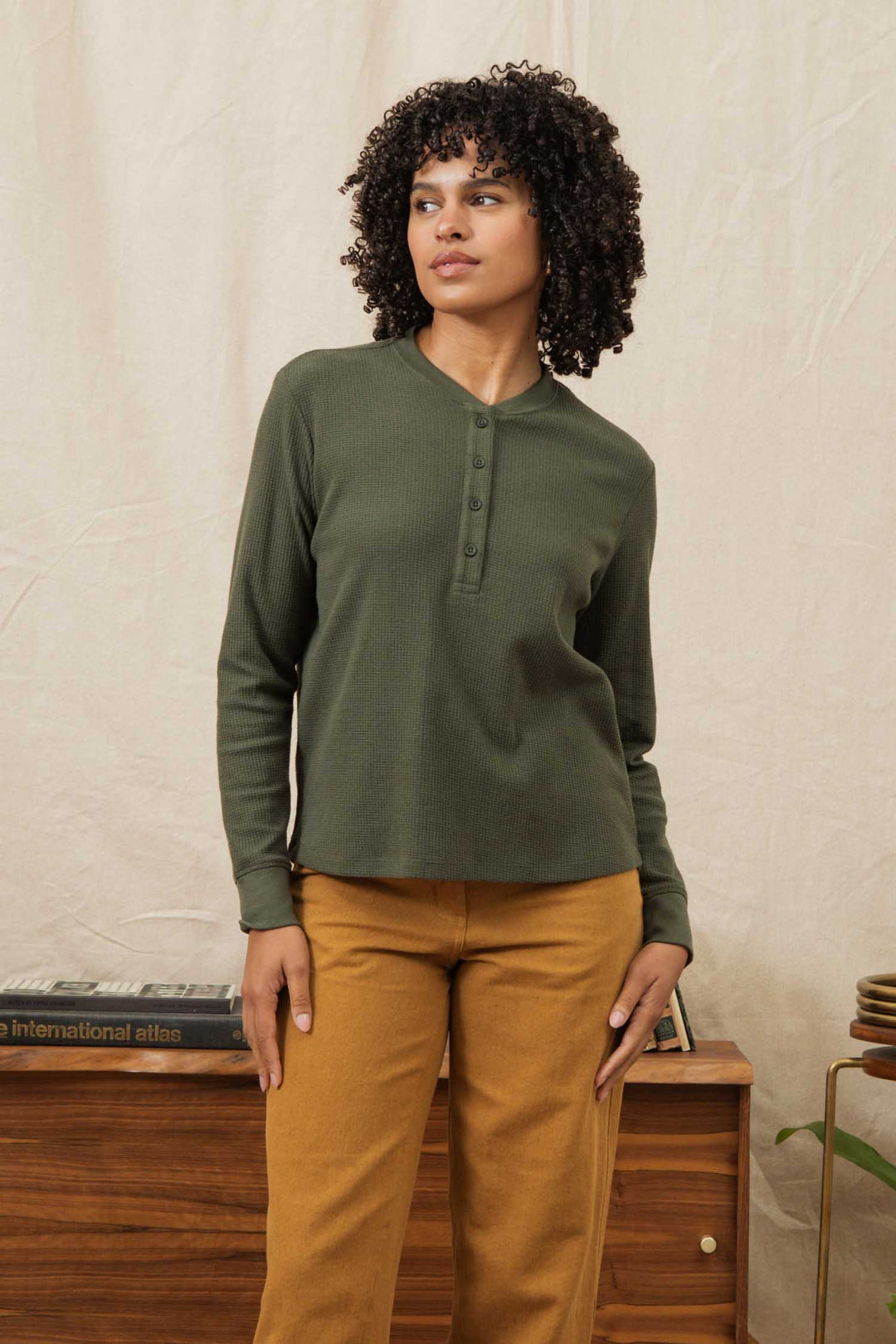 A woman with curly hair stands indoors by a beige curtain and wooden furniture, wearing the Bridge & Burn Women's Recycled Waffle Henley in Forest Melange with mustard yellow pants, looking to the side.