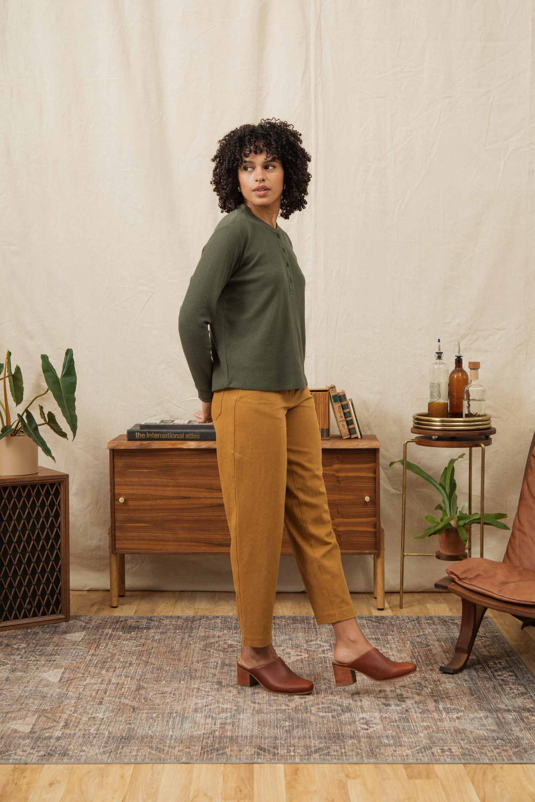 A woman with curly hair stands on a rug indoors, wearing the Bridge & Burn Women's Recycled Waffle Henley in Forest Melange, mustard pants, and brown shoes, posed before a wooden console surrounded by plants and decor.